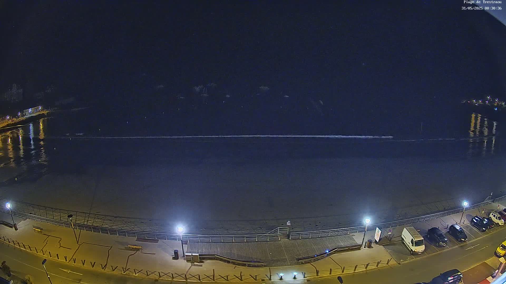 A nighttime view of a beach with calm water, a boardwalk, and a street lined with parked cars.