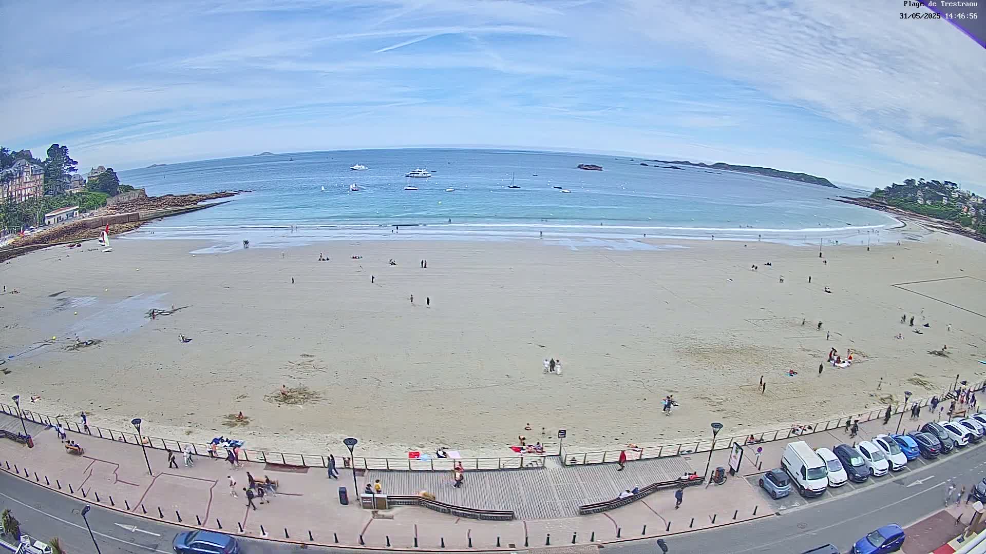 A mostly sunny day at a sandy beach with calm waves, several boats on the water, and people scattered across the sand and boardwalk.