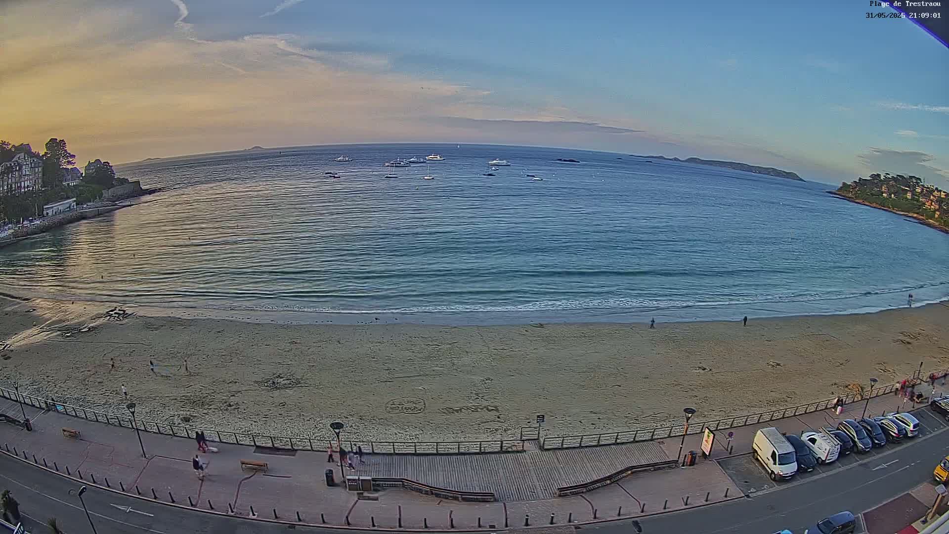 A sandy beach with a few people on it and several boats on calm water, under a mostly clear sunset sky.