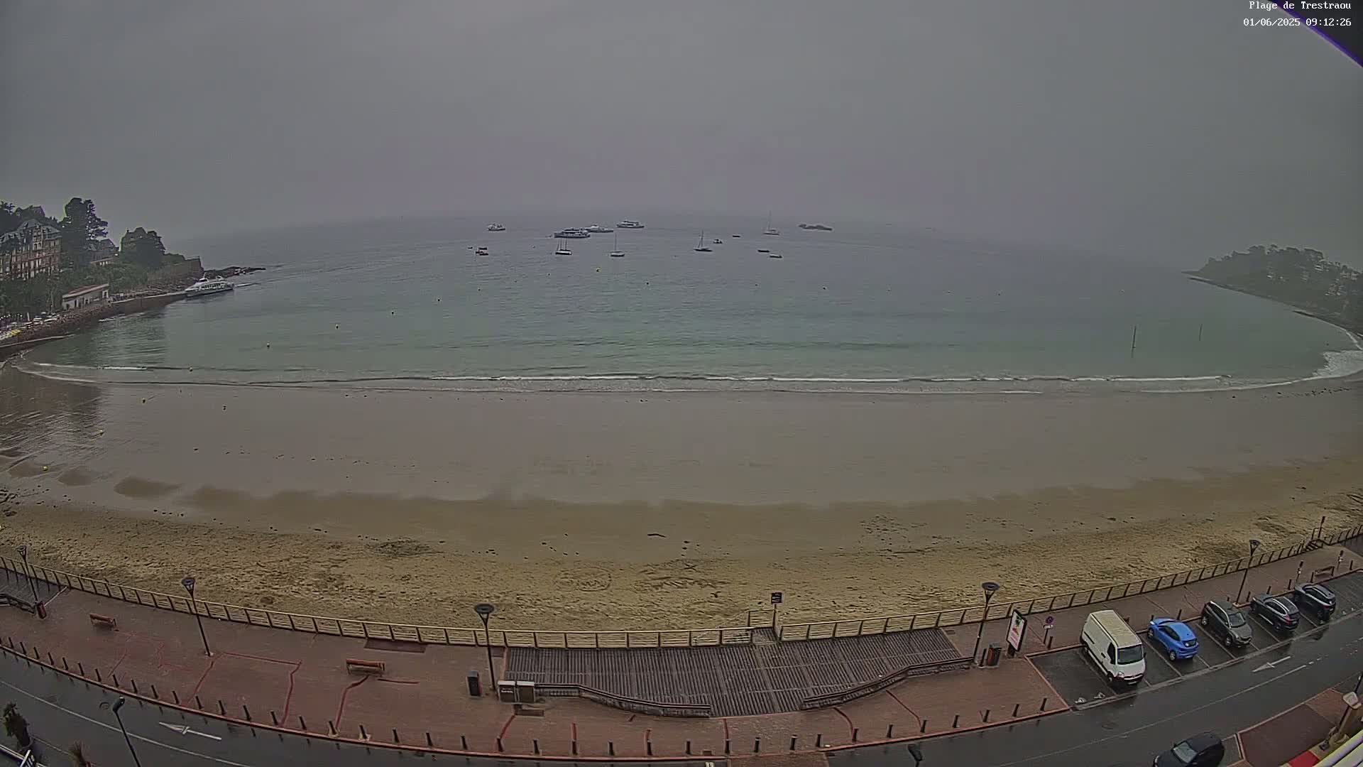 A sandy beach curves into a calm, foggy bay with several boats anchored offshore; a paved walkway and a parking lot with cars are visible in the foreground.