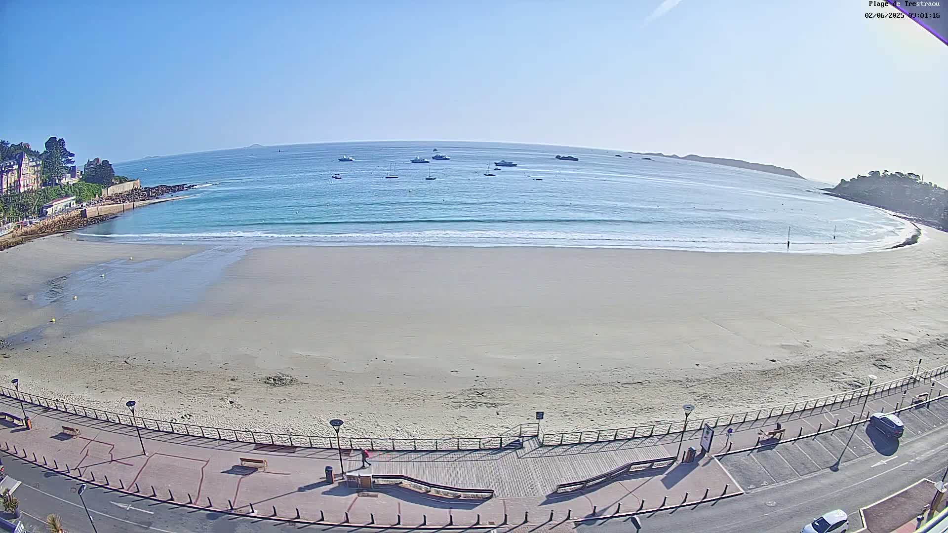 A wide sandy beach with gentle waves under a clear blue sky, bordered by a paved walkway and buildings, with several boats visible in the distance.