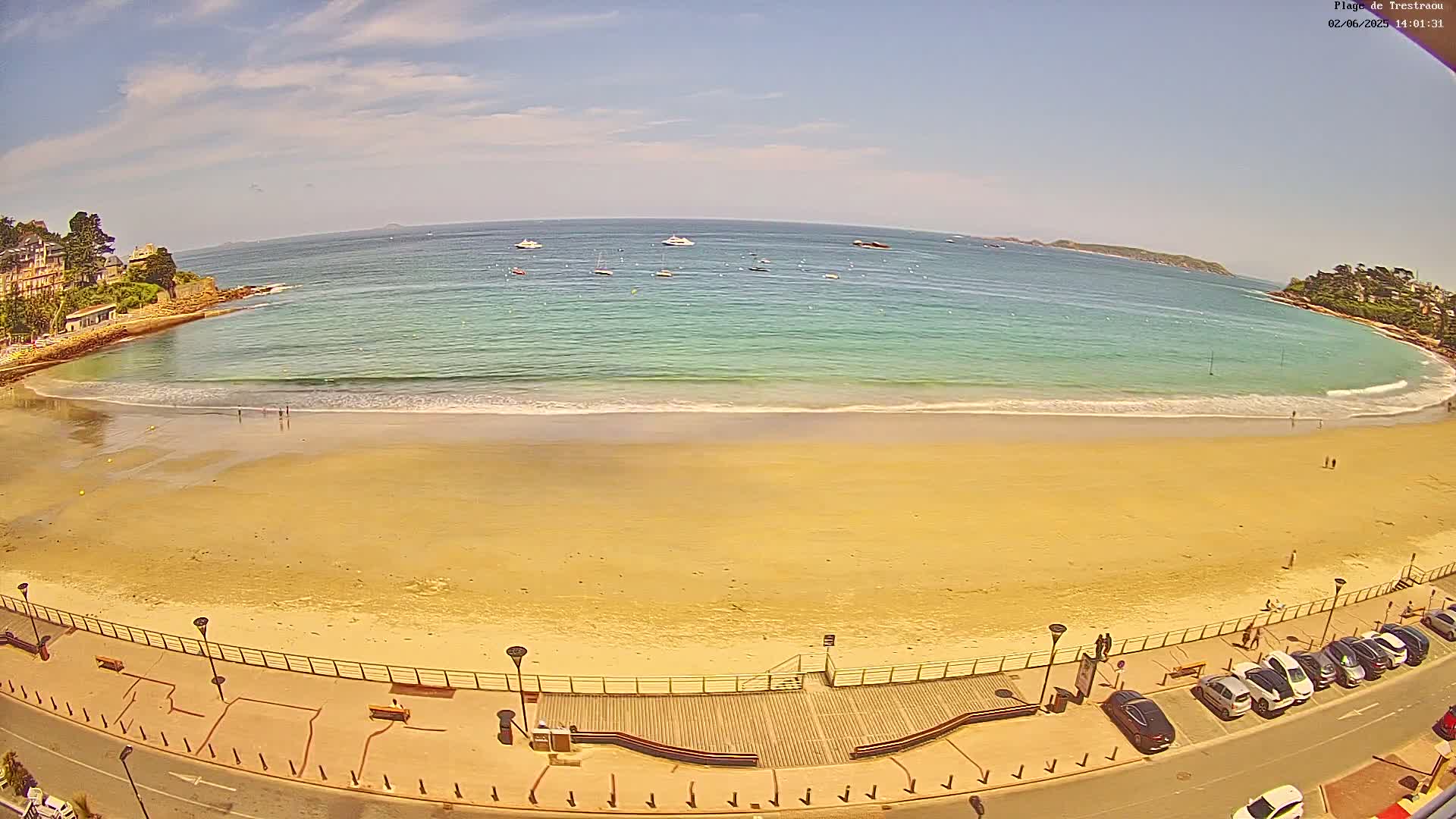 A sandy beach with calm, turquoise water stretches beneath a mostly sunny sky, with a few boats visible in the distance.