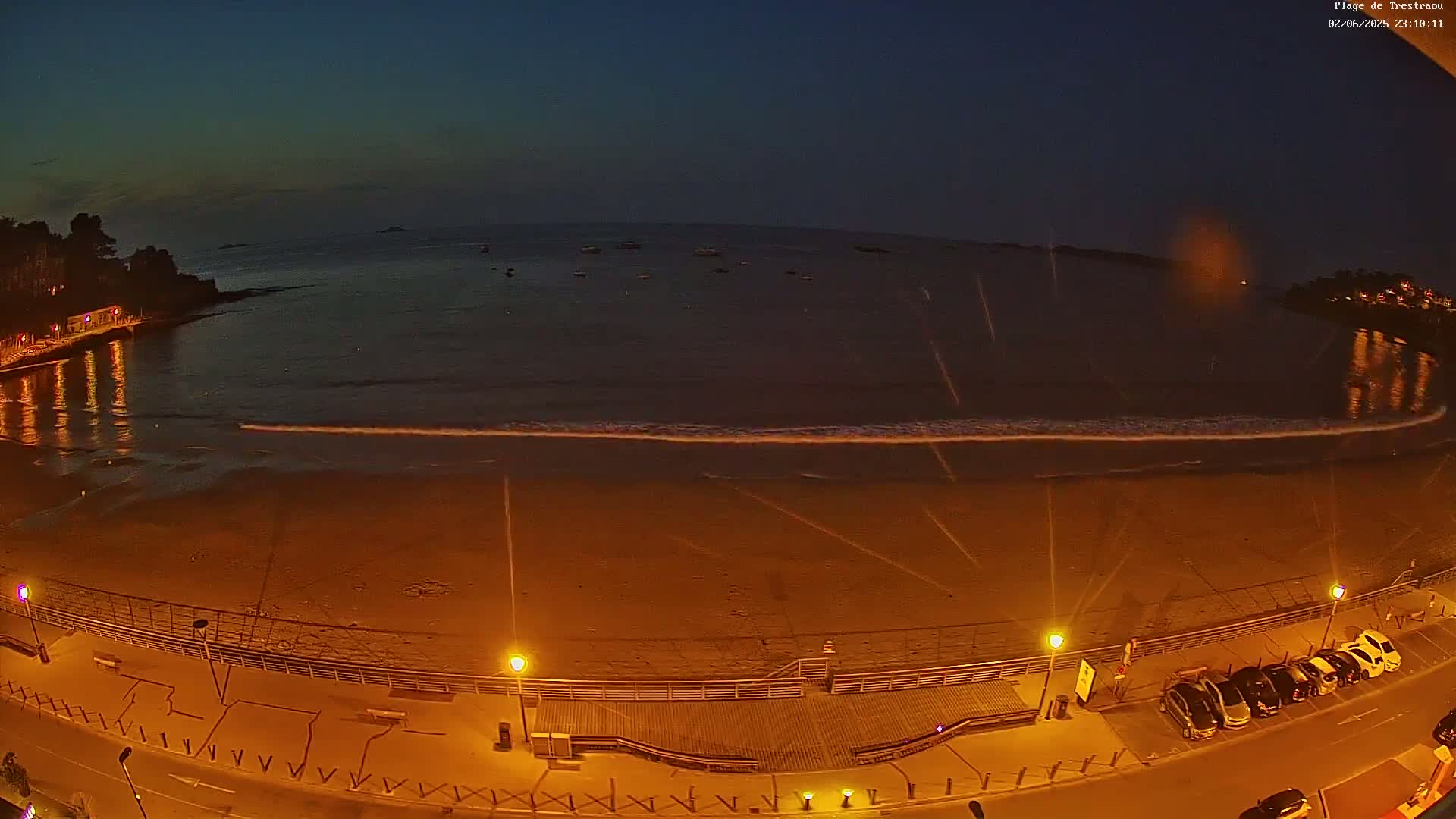A dark, nighttime beach scene shows a sandy shore with gently lapping waves under a clear, dark sky; the beach is illuminated by streetlights, and cars are parked along a roadway beside it.