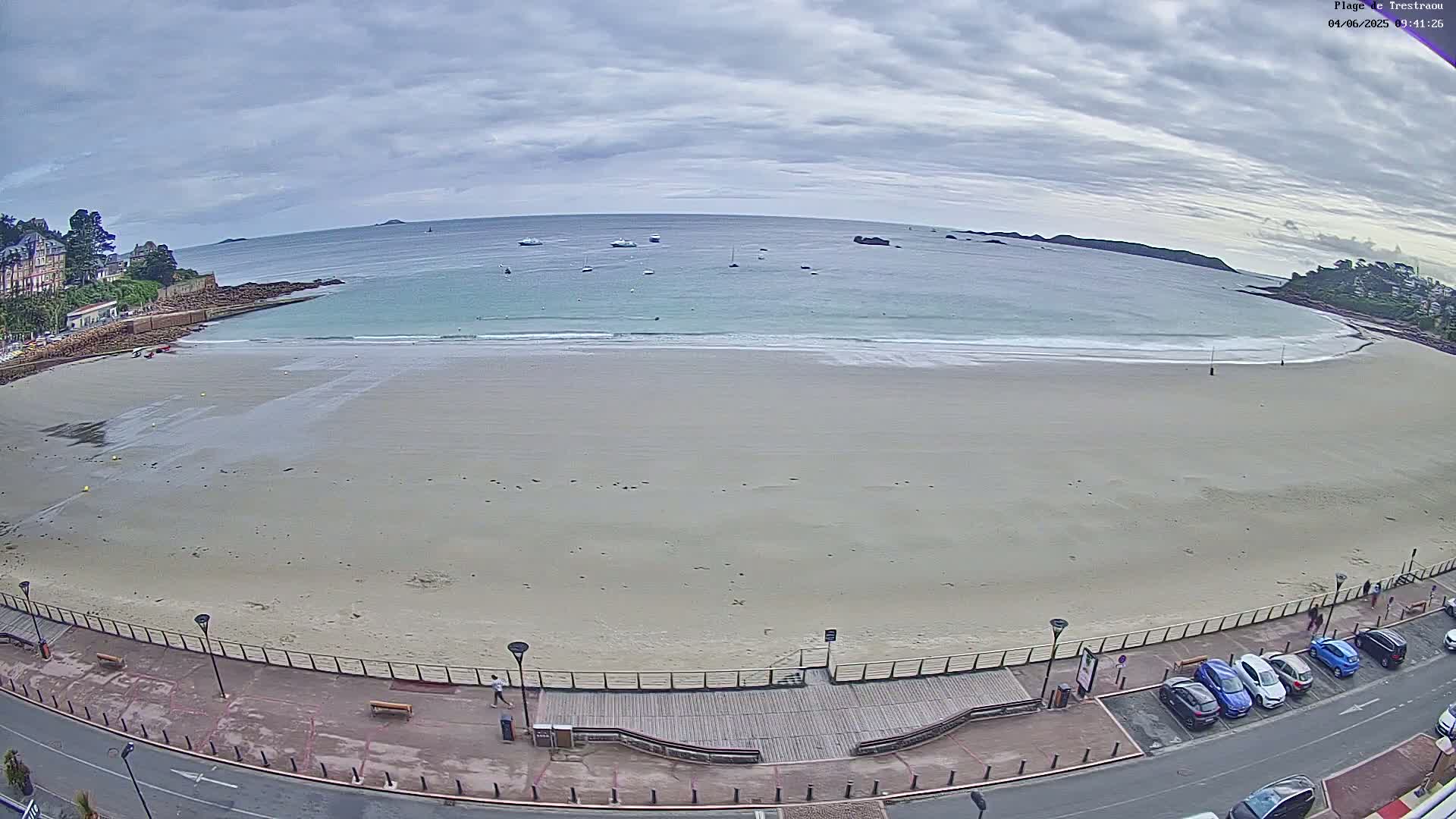 A mostly empty sandy beach with calm, light-blue water under a cloudy sky, bordered by a paved walkway and a row of parked cars.