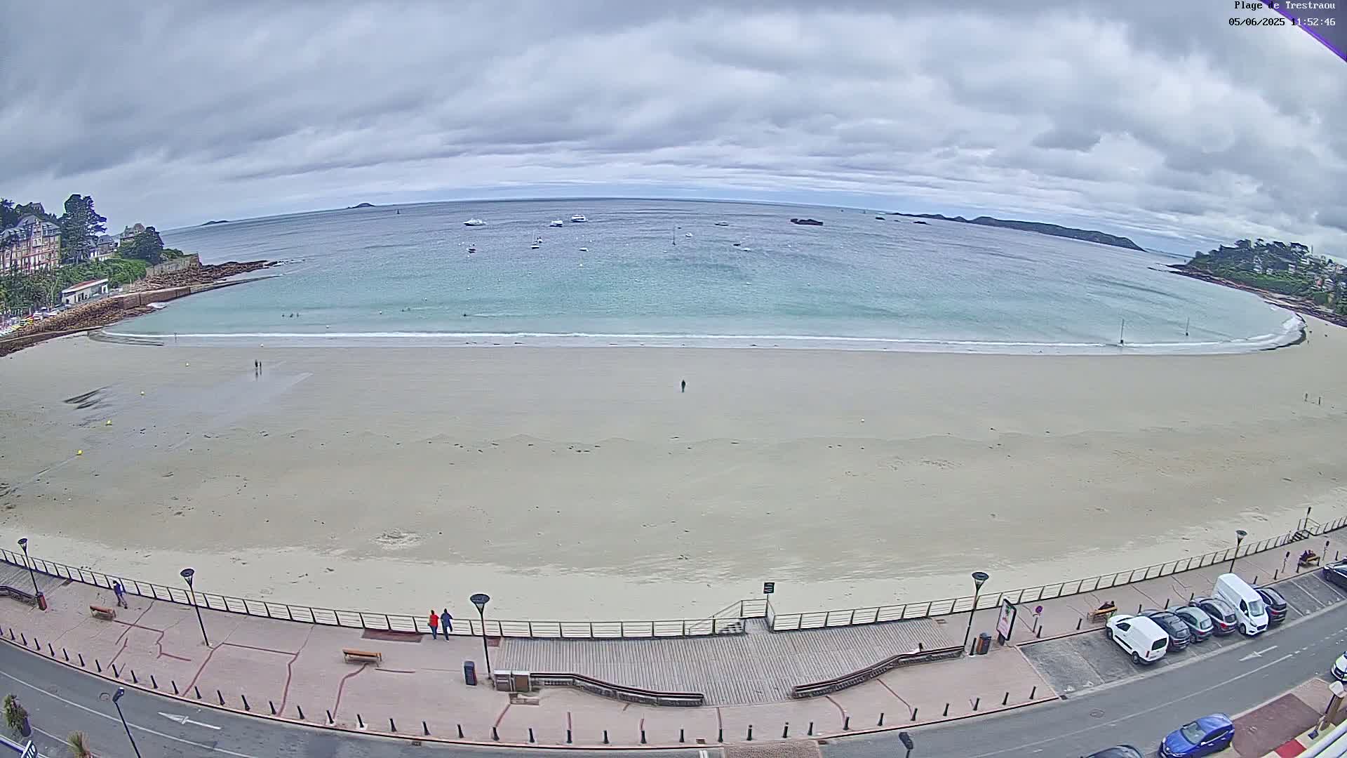 A wide, sandy beach stretches along a calm, light-blue ocean under an overcast sky, with a few people on the beach and cars parked along a road overlooking the water.