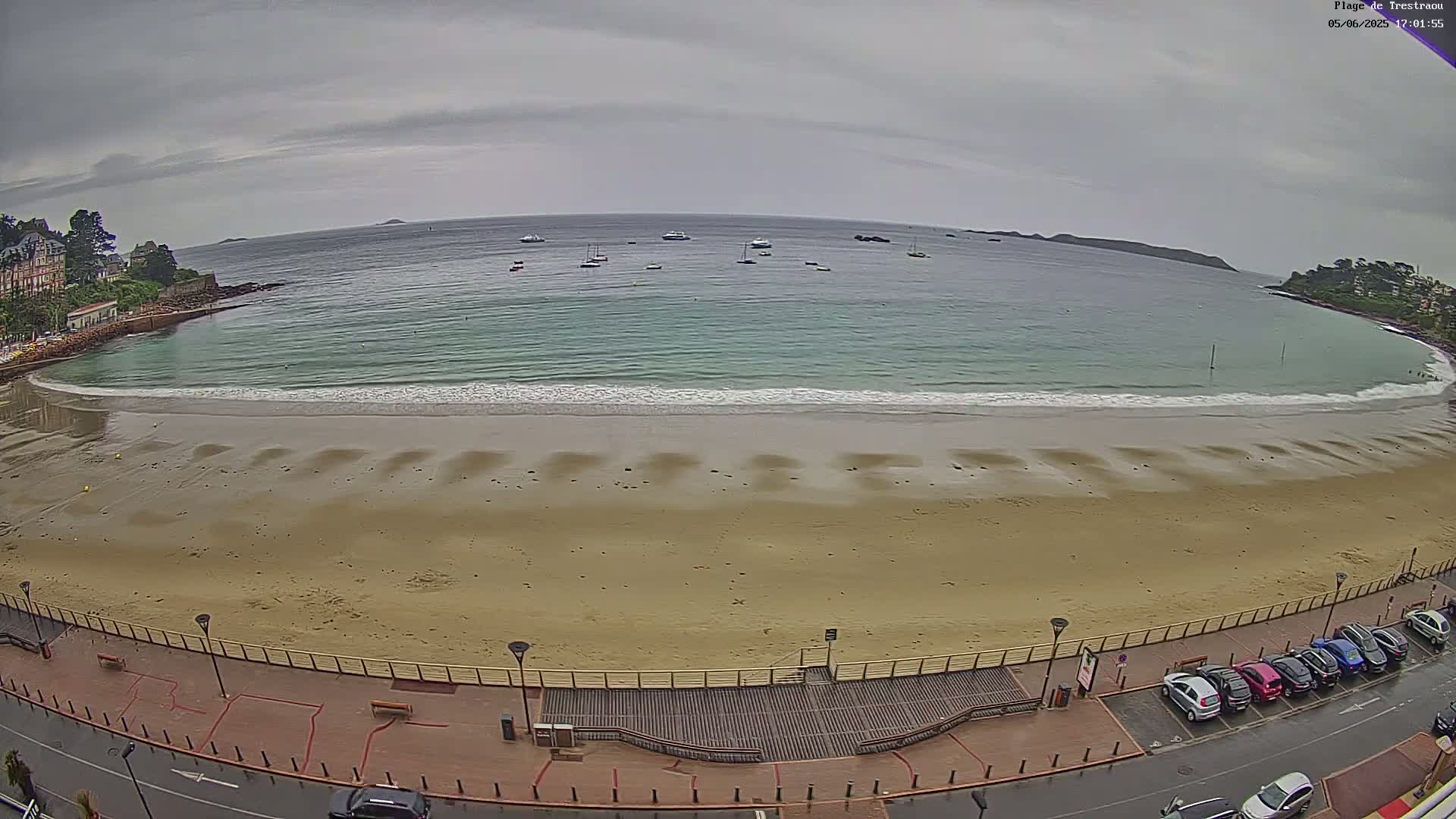 A sandy beach with gentle waves laps the shore under an overcast sky, with several boats visible in the calm water beyond.