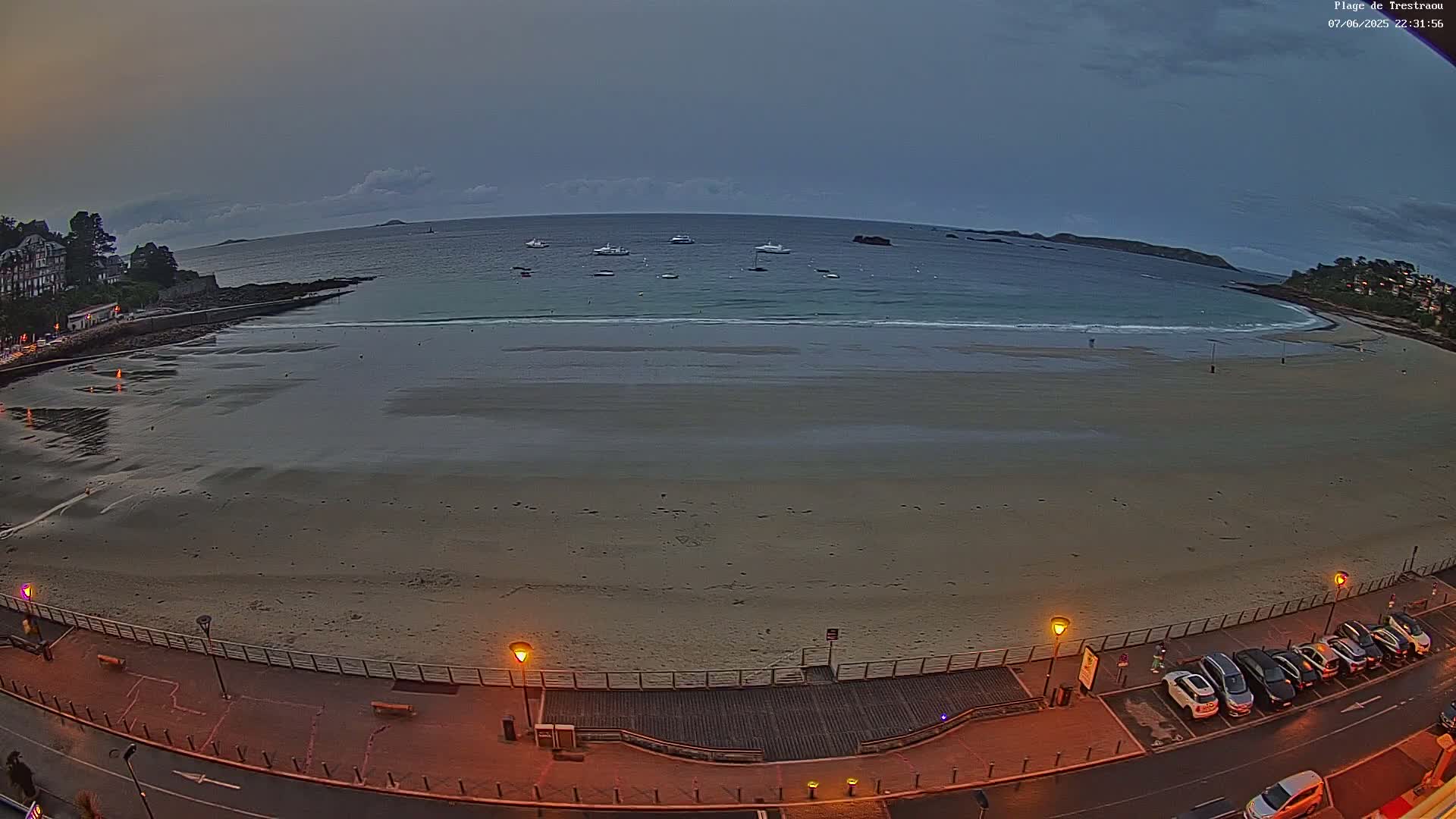 A long, sandy beach at dusk, with several boats visible in the calm water beyond, is illuminated by streetlights along a paved walkway next to a parking lot.
