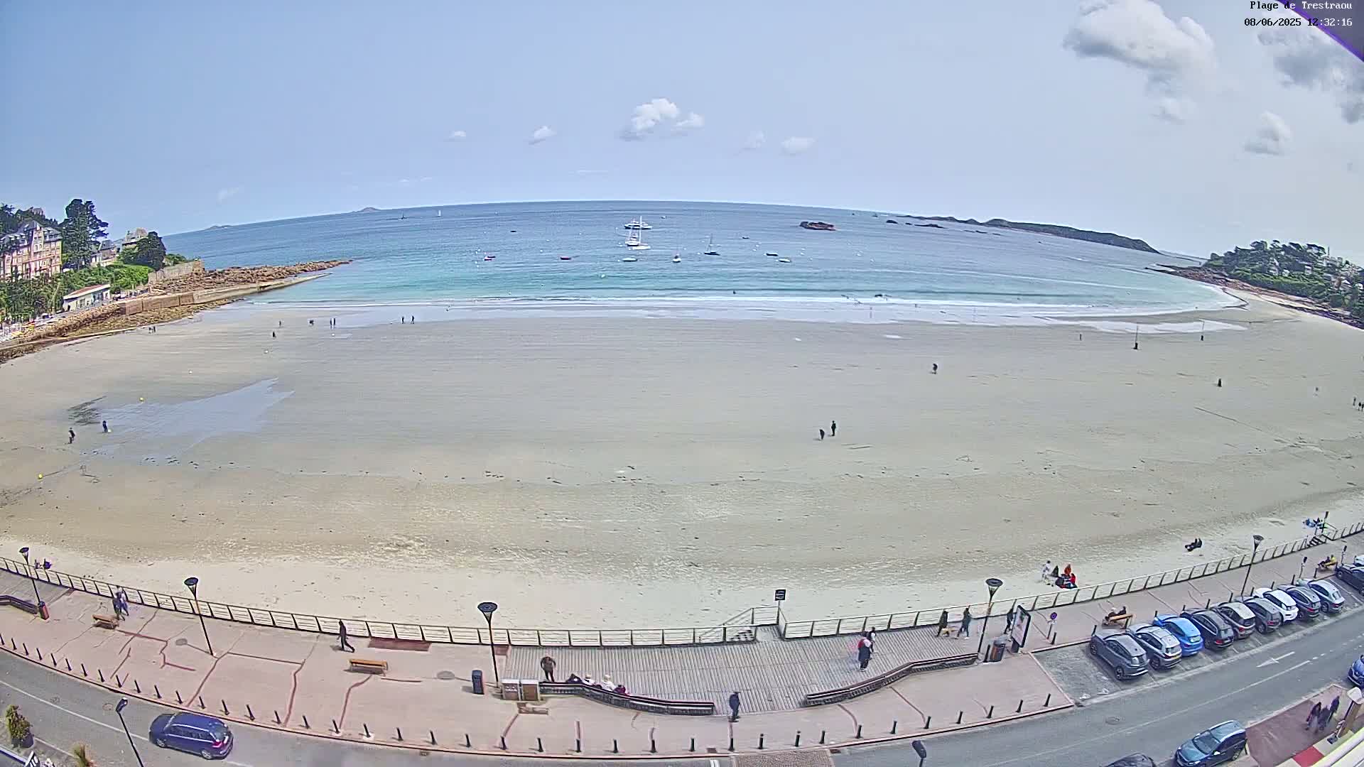 A sandy beach with a few people on it stretches along a calm, light-blue ocean under a mostly clear sky;  cars are parked along a road next to a boardwalk that runs the length of the beach.