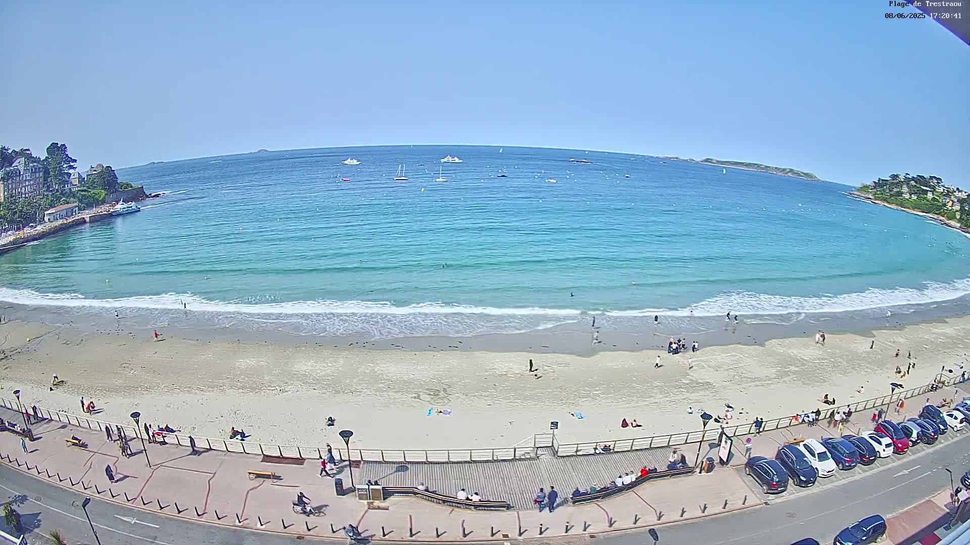 A sandy beach with light waves and numerous people enjoying a sunny day, bordered by a boardwalk with parked cars and buildings in the background.