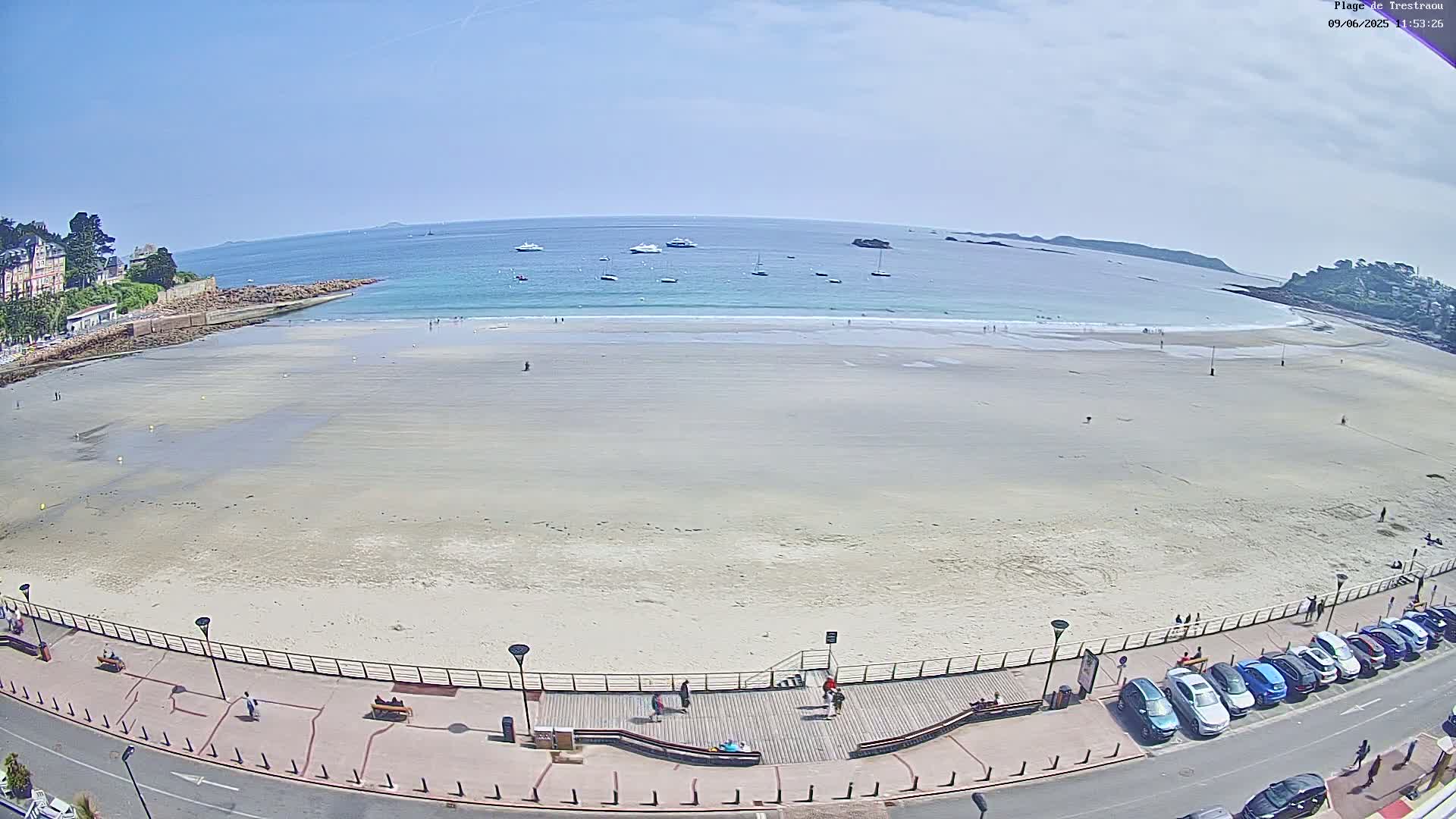 A wide sandy beach stretches out to a calm ocean with several boats visible on the water under a clear blue sky; a boardwalk and parked cars line the beach.