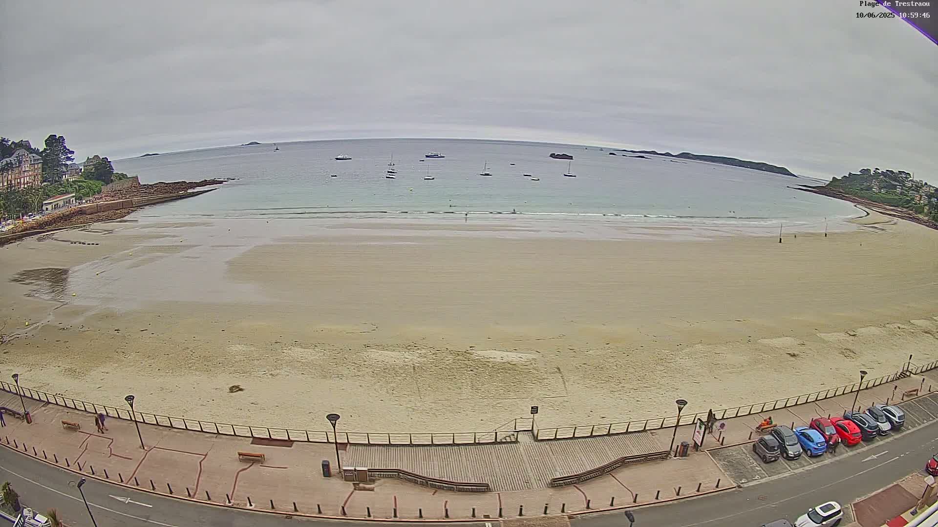 A sandy beach with a few people and several cars parked along a boardwalk overlooks a calm, overcast ocean with several boats.