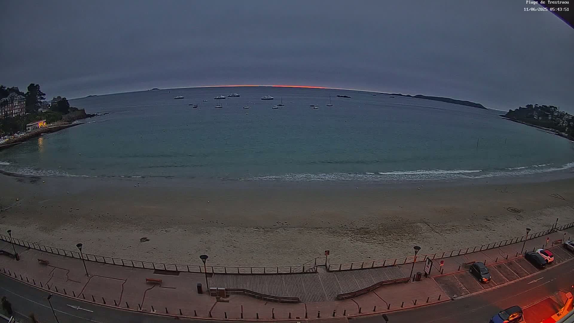 A sandy beach with a boardwalk and parked cars is shown at dawn, with calm waves lapping the shore and several boats visible on the water in the distance.