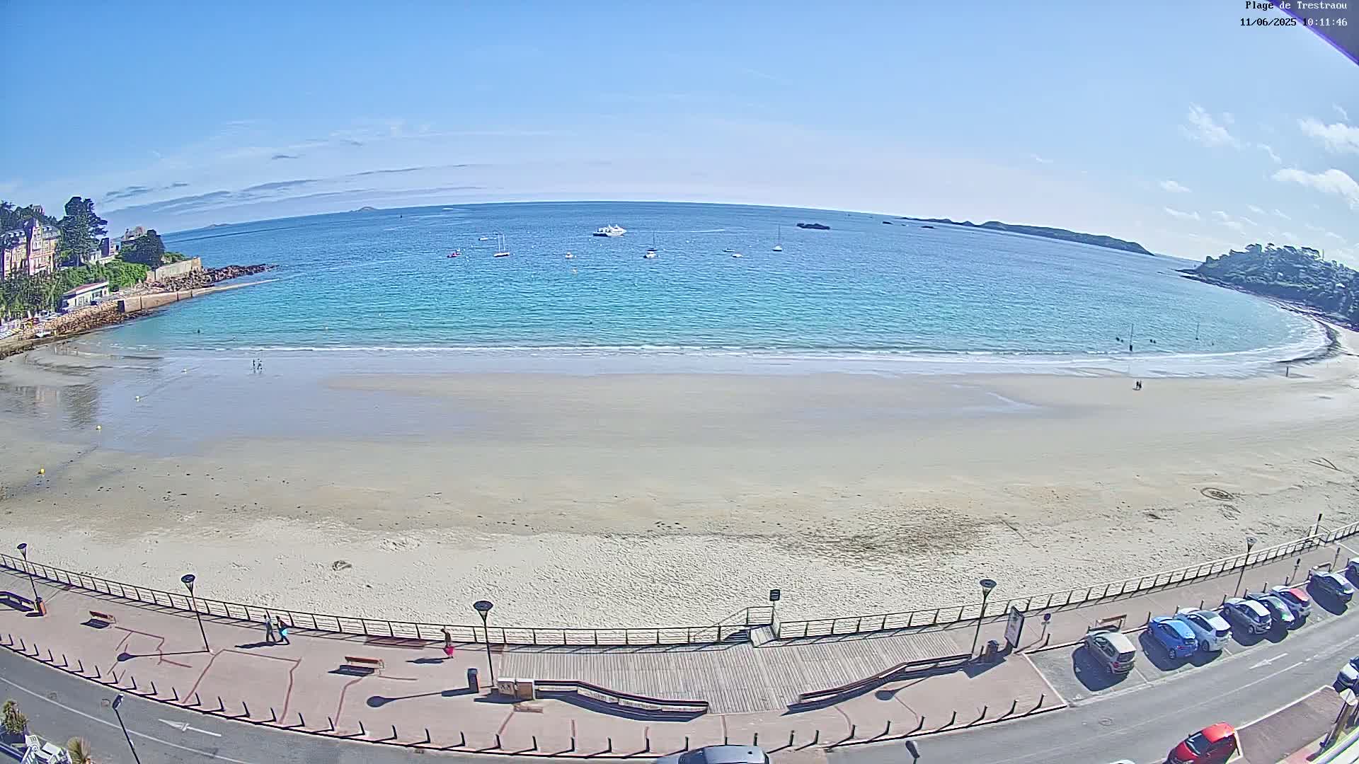 A sandy beach with calm, turquoise water under a clear blue sky is bordered by a paved walkway lined with streetlights and parked cars.