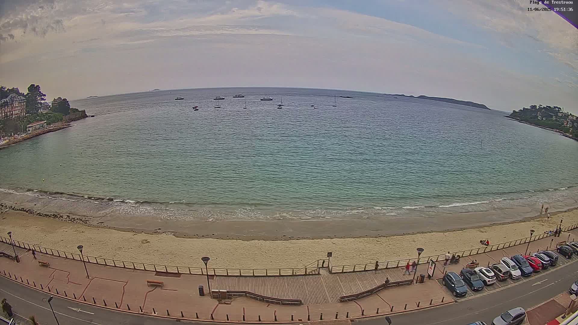 A sandy beach with calm, light-blue water and several boats in the distance under a partly cloudy sky.