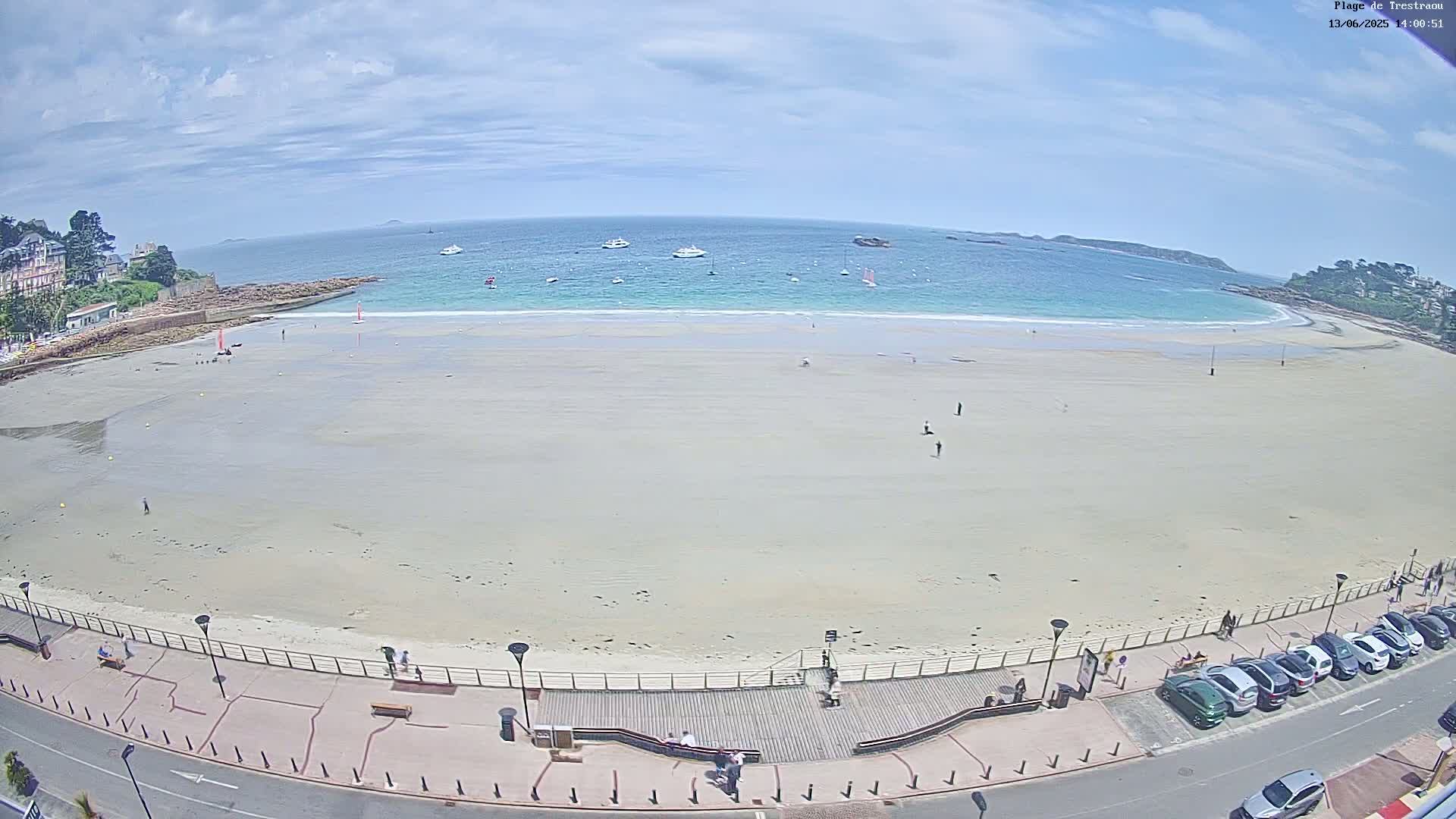 A wide, sandy beach stretches along a turquoise ocean under a partly cloudy sky, with several small boats visible on the water and a few people scattered on the sand and boardwalk.