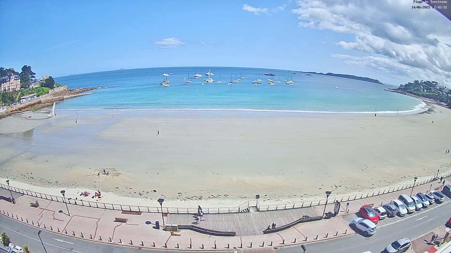 A sandy beach with calm, turquoise water and several boats in the distance under a partly cloudy sky.