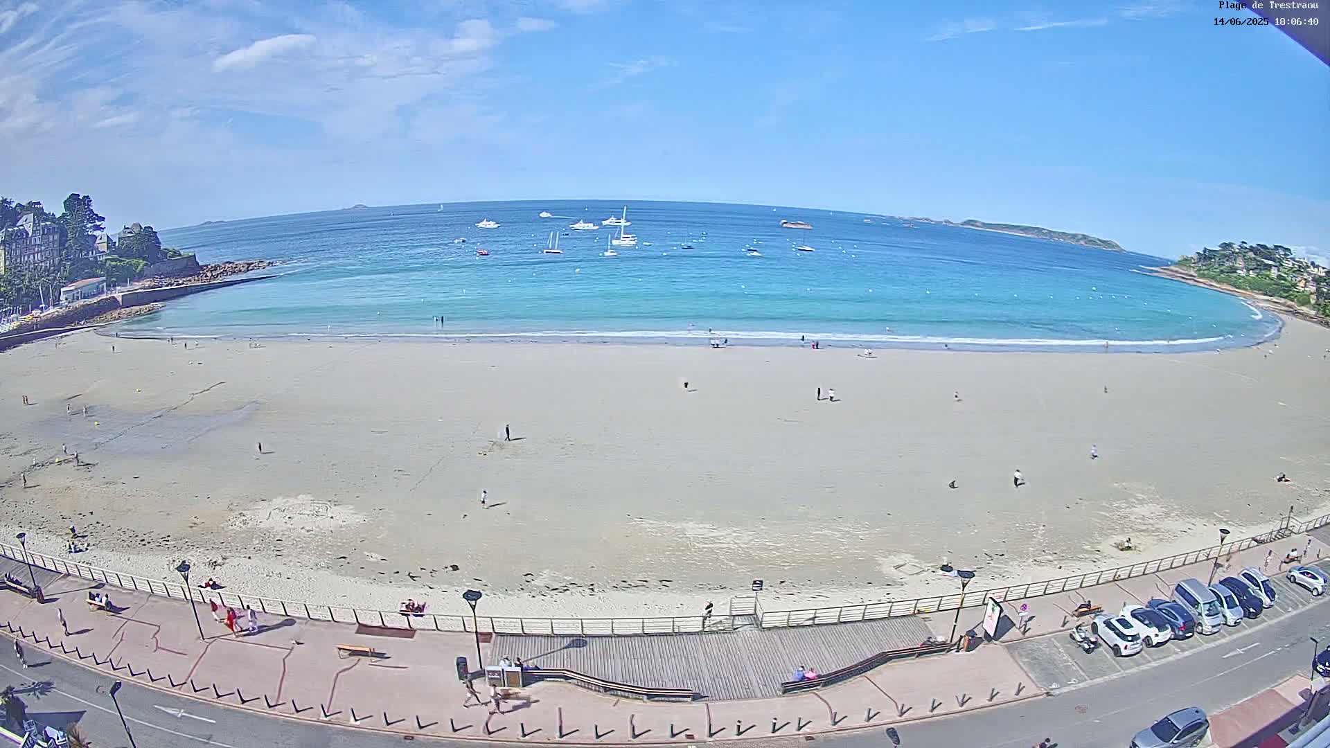 A sandy beach with a few people on it curves gently into a calm, turquoise ocean dotted with sailboats under a mostly sunny sky.
