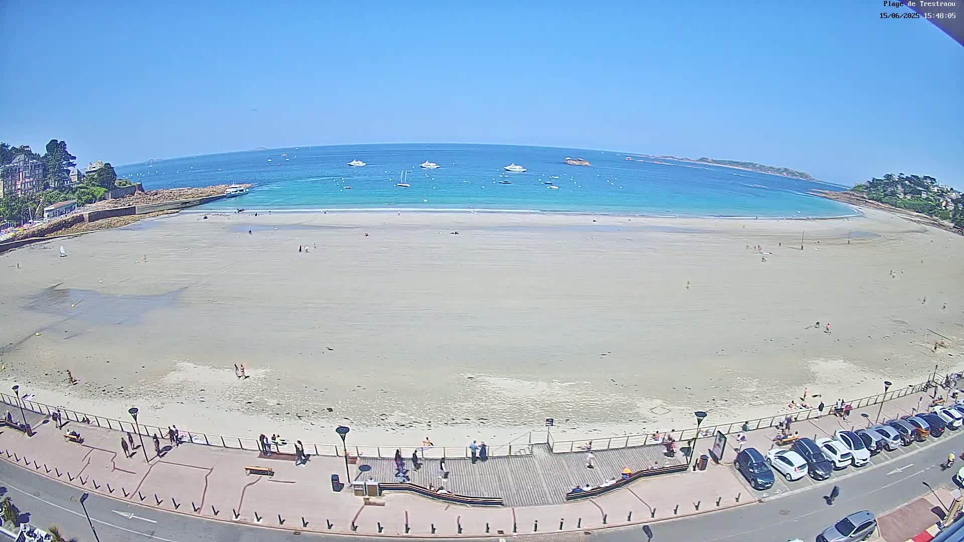 A wide, sandy beach stretches along a turquoise ocean under a clear blue sky, with several boats visible offshore and people scattered along the shore and boardwalk.