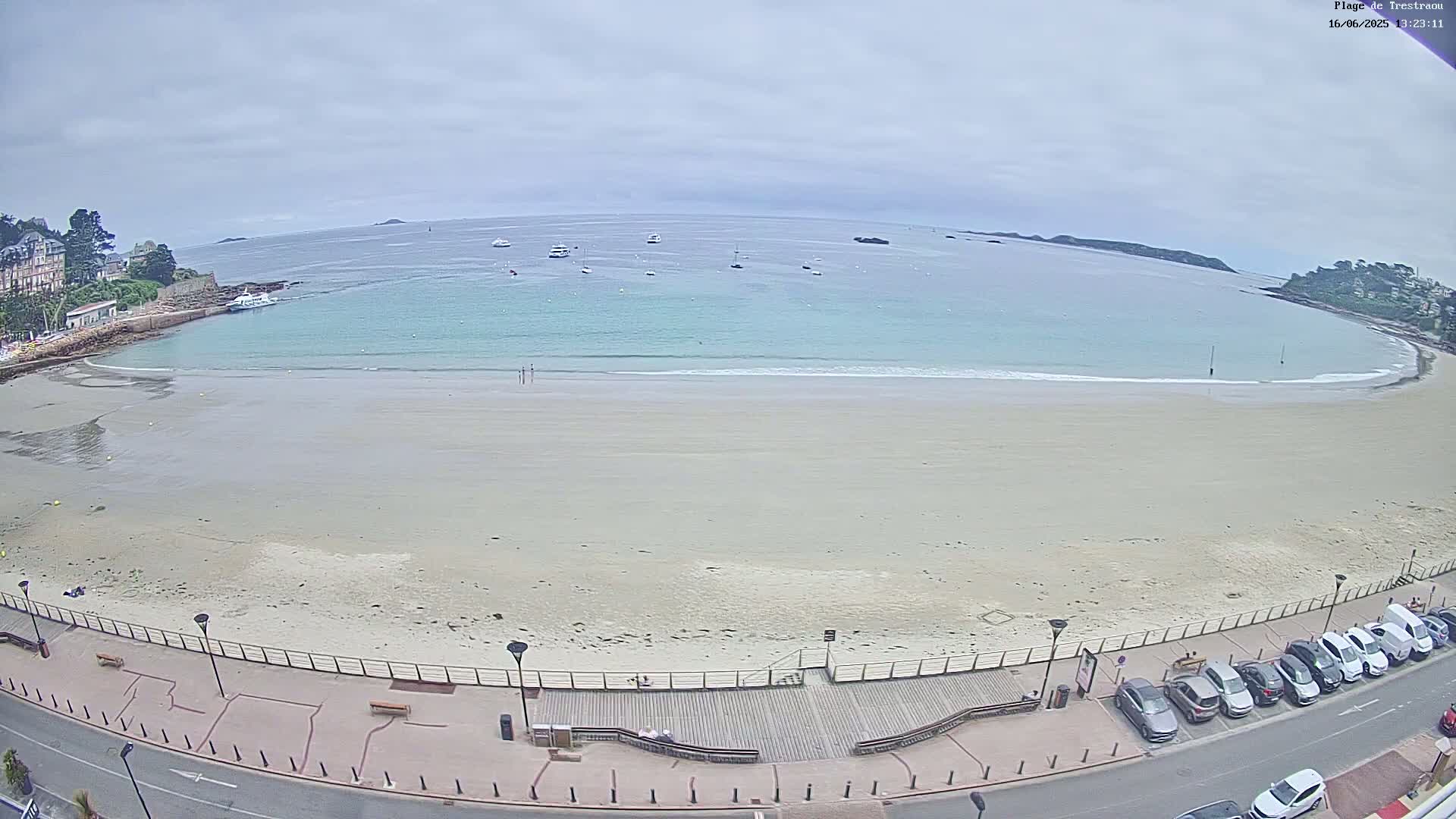 An expansive sandy beach with a calm, light-blue ocean under an overcast sky is viewed from an elevated position, showing a boardwalk, parked cars, and buildings in the distance.