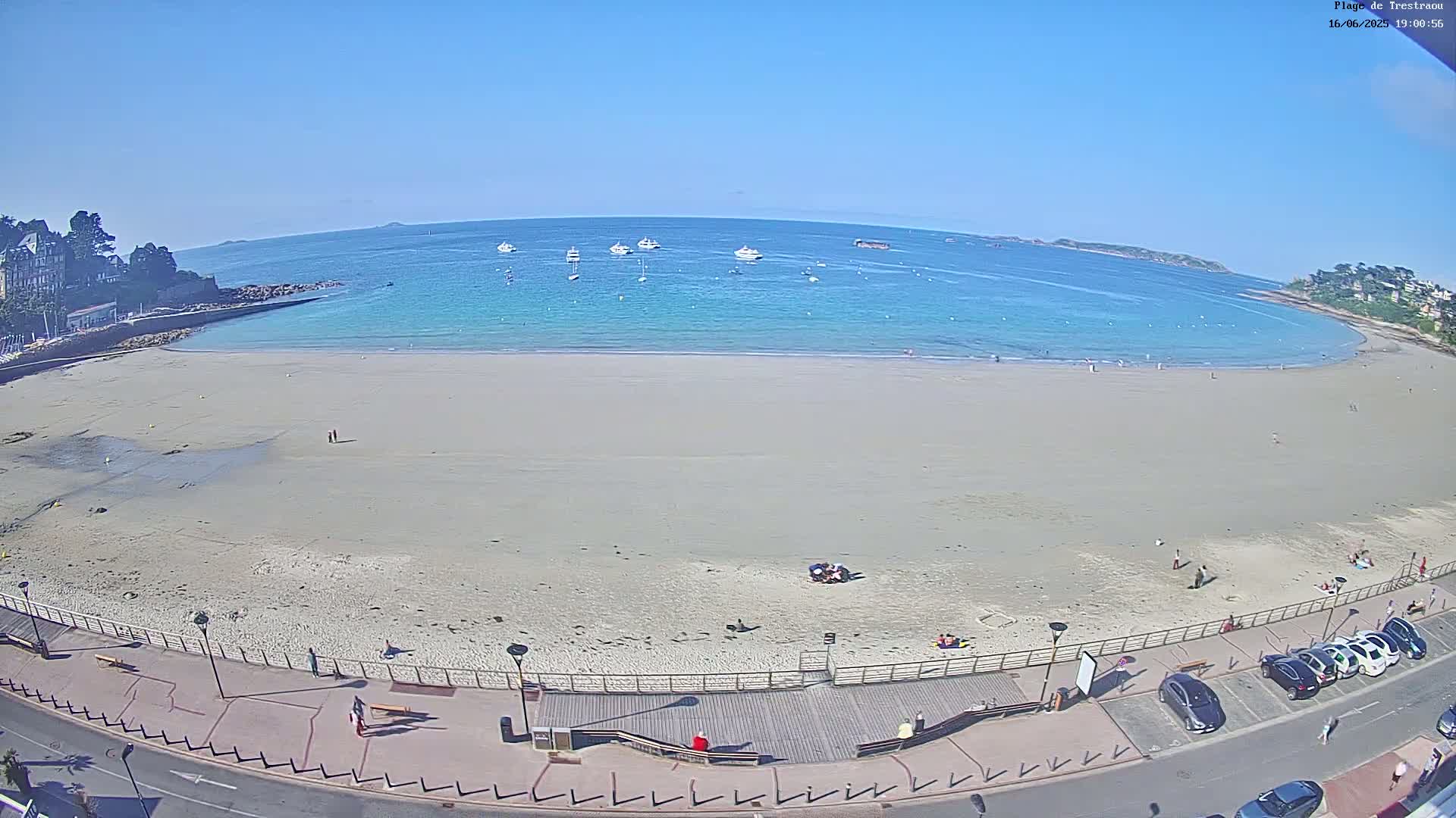 A wide sandy beach stretches along a calm, turquoise ocean under a clear blue sky, with several boats visible in the water and people scattered on the sand and boardwalk.