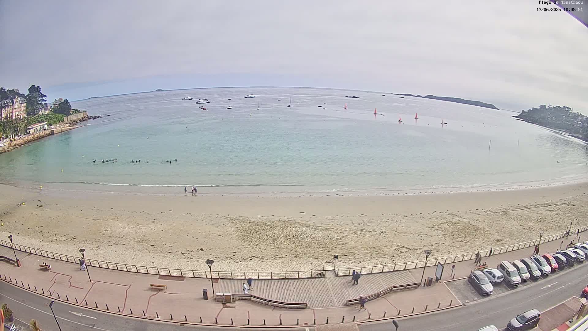 A sandy beach with several people on it, a calm, light-blue body of water with sailboats and people swimming, and a parking lot with cars next to it under an overcast sky.