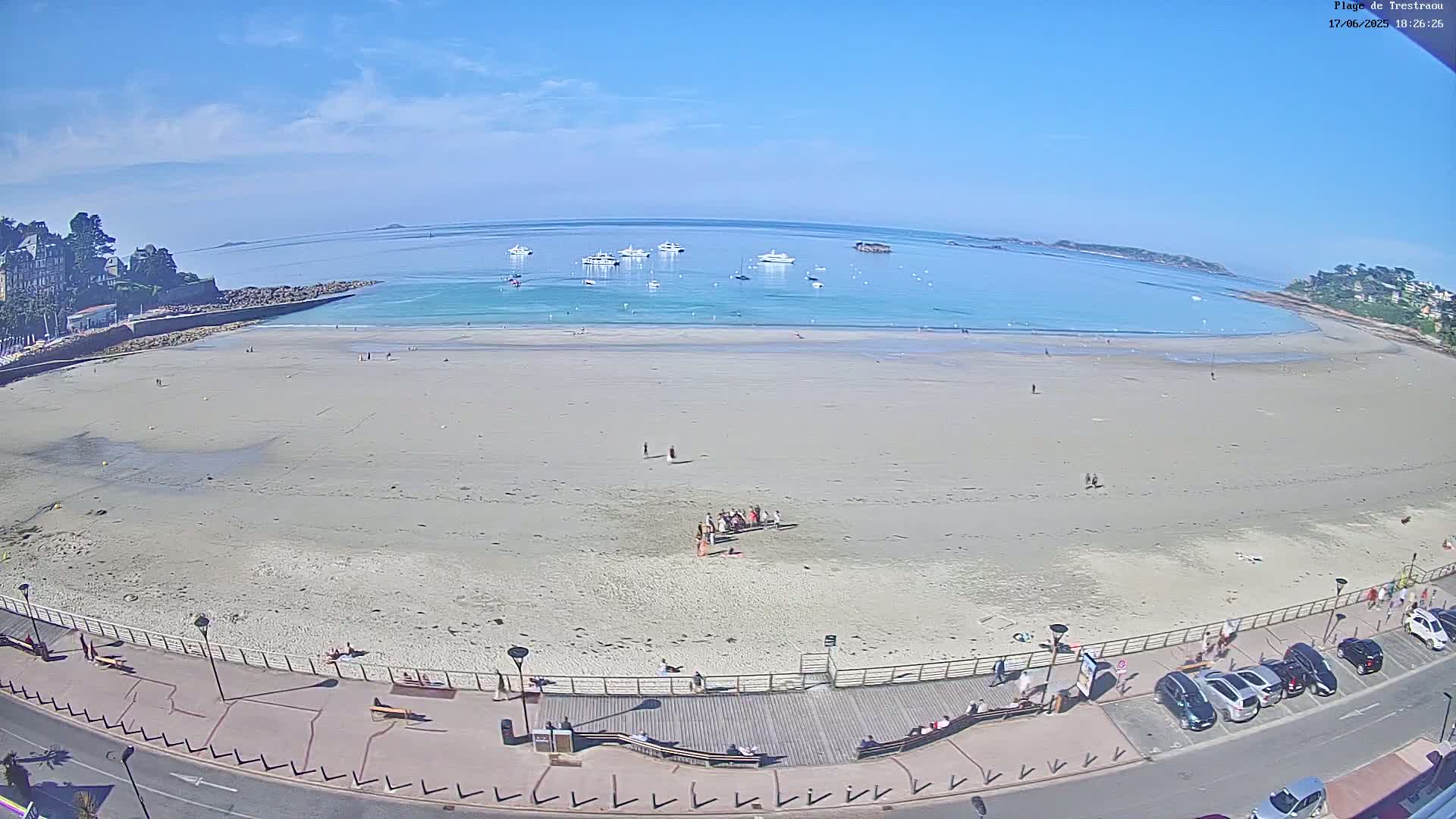 A wide sandy beach with a few people on it, several boats in the calm blue water, and buildings along the shoreline under a clear sunny sky.