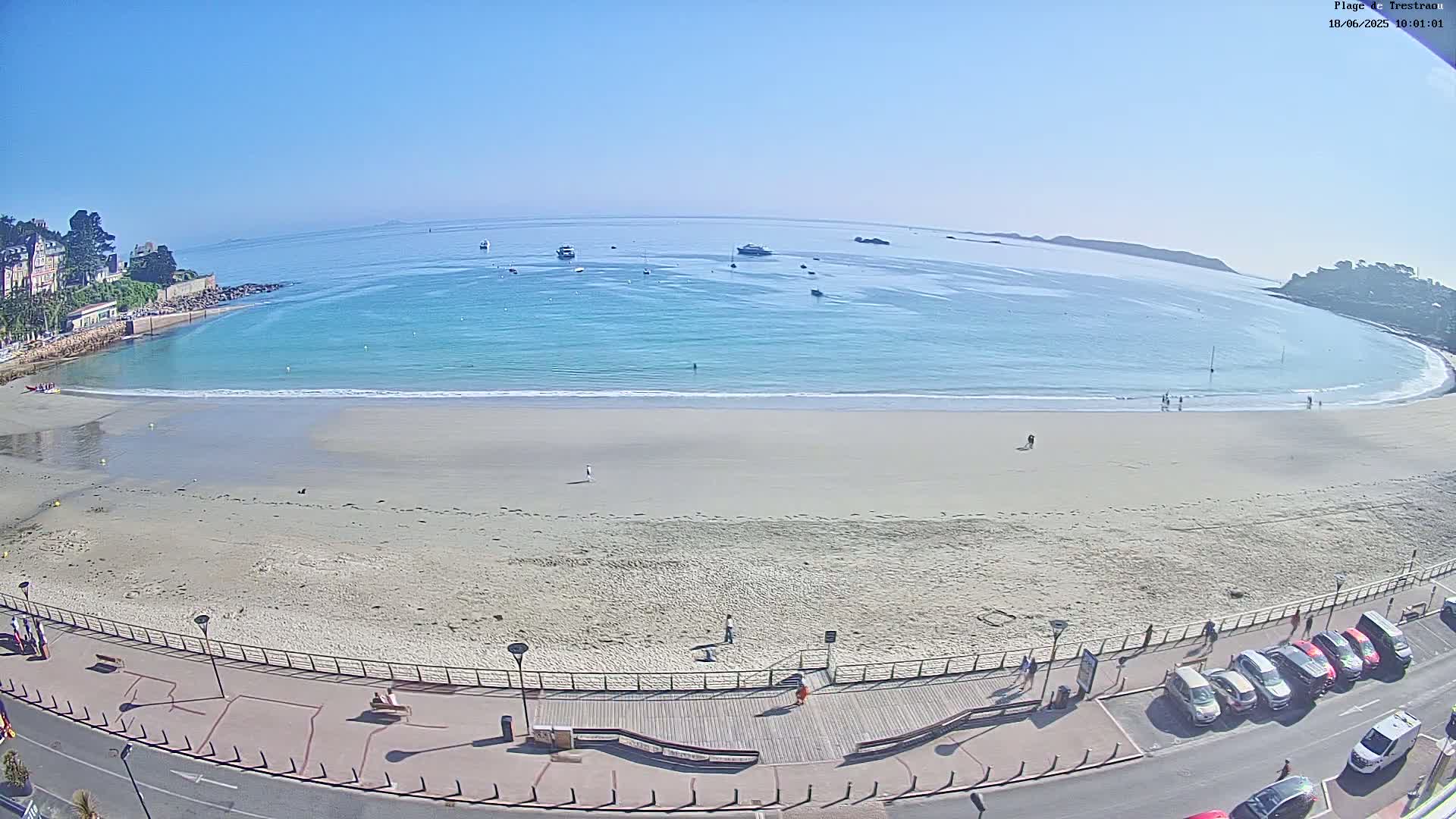 A sandy beach curves around a calm, light-blue bay on a sunny day, with several boats visible in the water and cars parked along the road adjacent to the beach.
