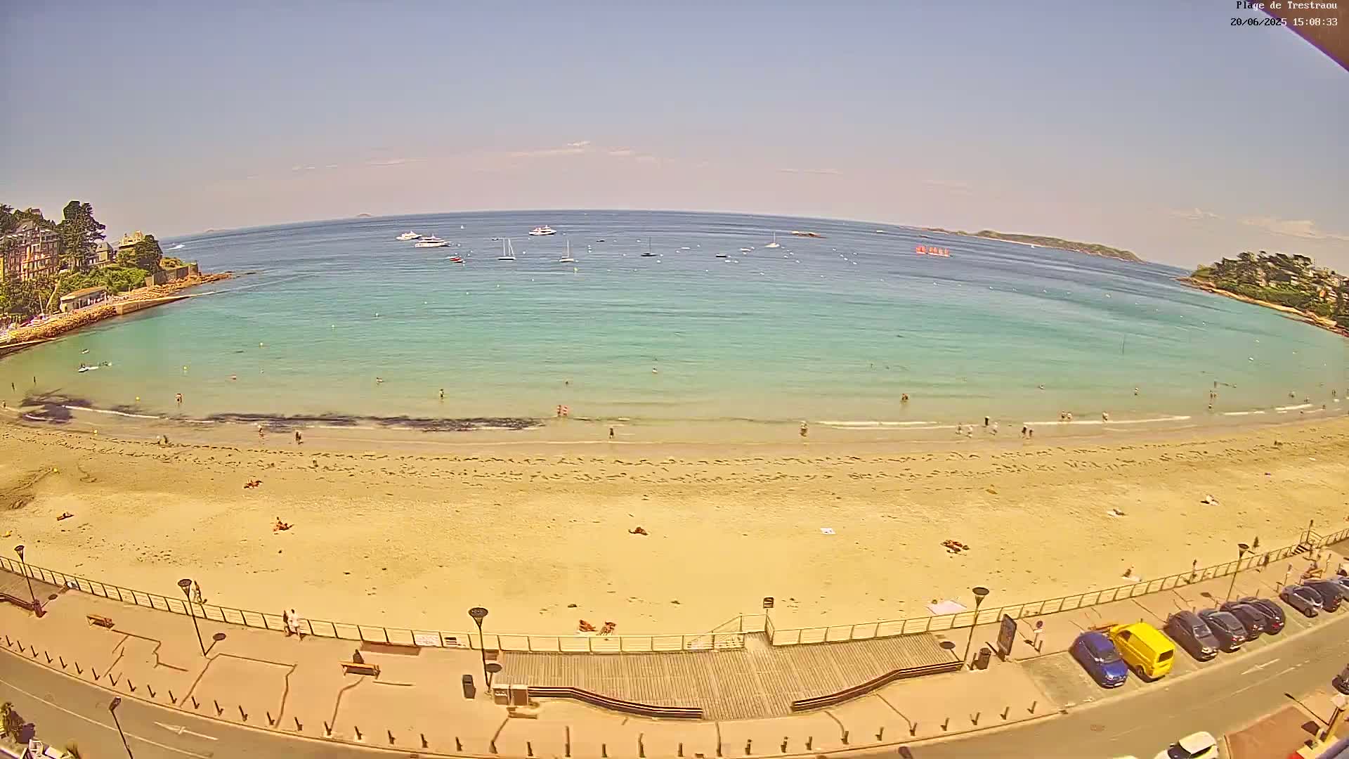 A sandy beach with a few people on it curves around a calm, turquoise bay on a sunny day, with several boats visible in the distance.