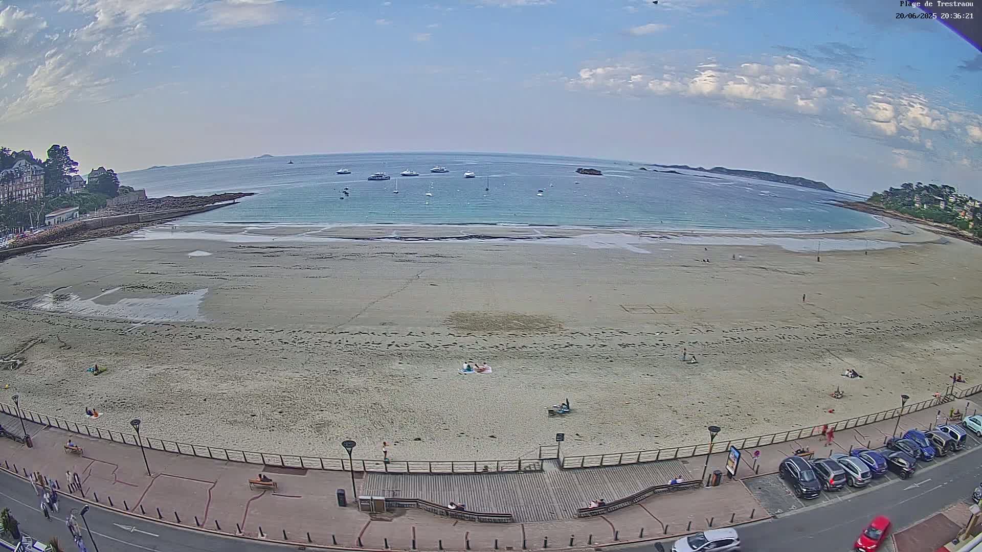 A wide sandy beach with a few people on it, bordered by a roadway and buildings, under a partly cloudy sky.
