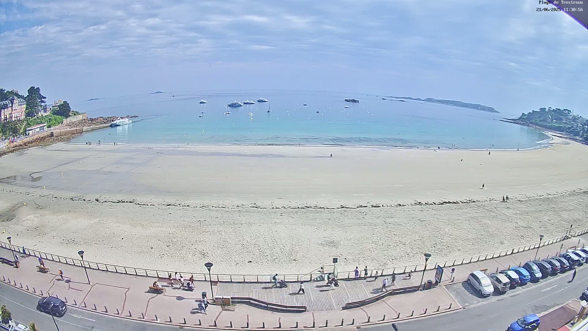 A wide sandy beach with calm, clear turquoise water, several boats in the distance, and a few people walking along the boardwalk under sunny skies.