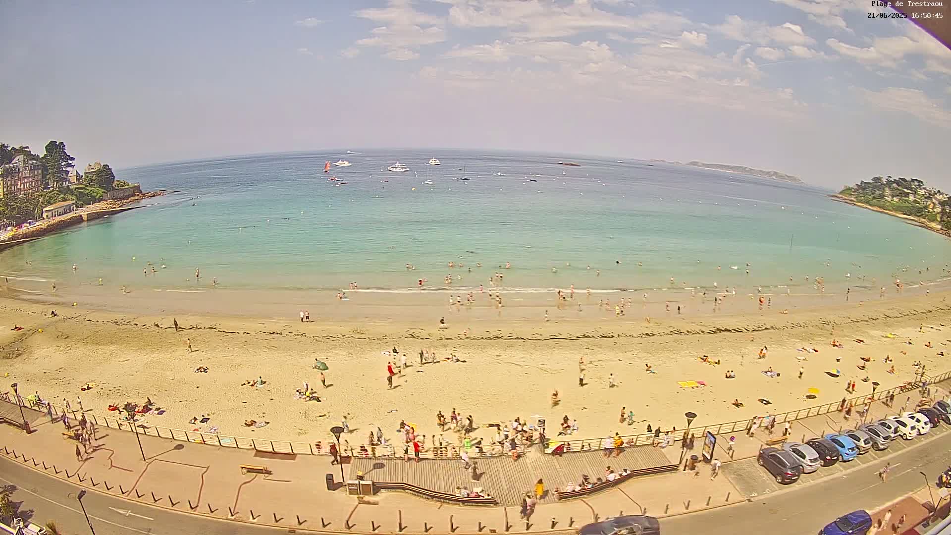 A sandy beach with many people swimming and sunbathing, bordered by a promenade with parked cars, all under a partly cloudy sky.