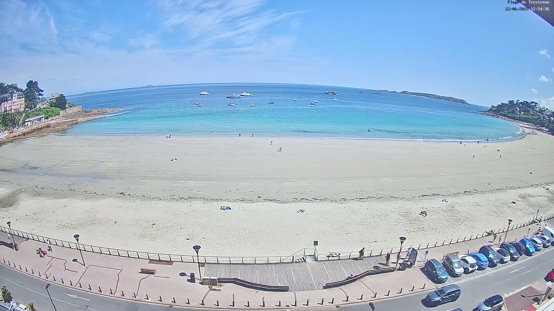 A sandy beach with a few people on it, bordered by a boardwalk and parking lot, stretches along a calm, turquoise ocean under a clear blue sky.