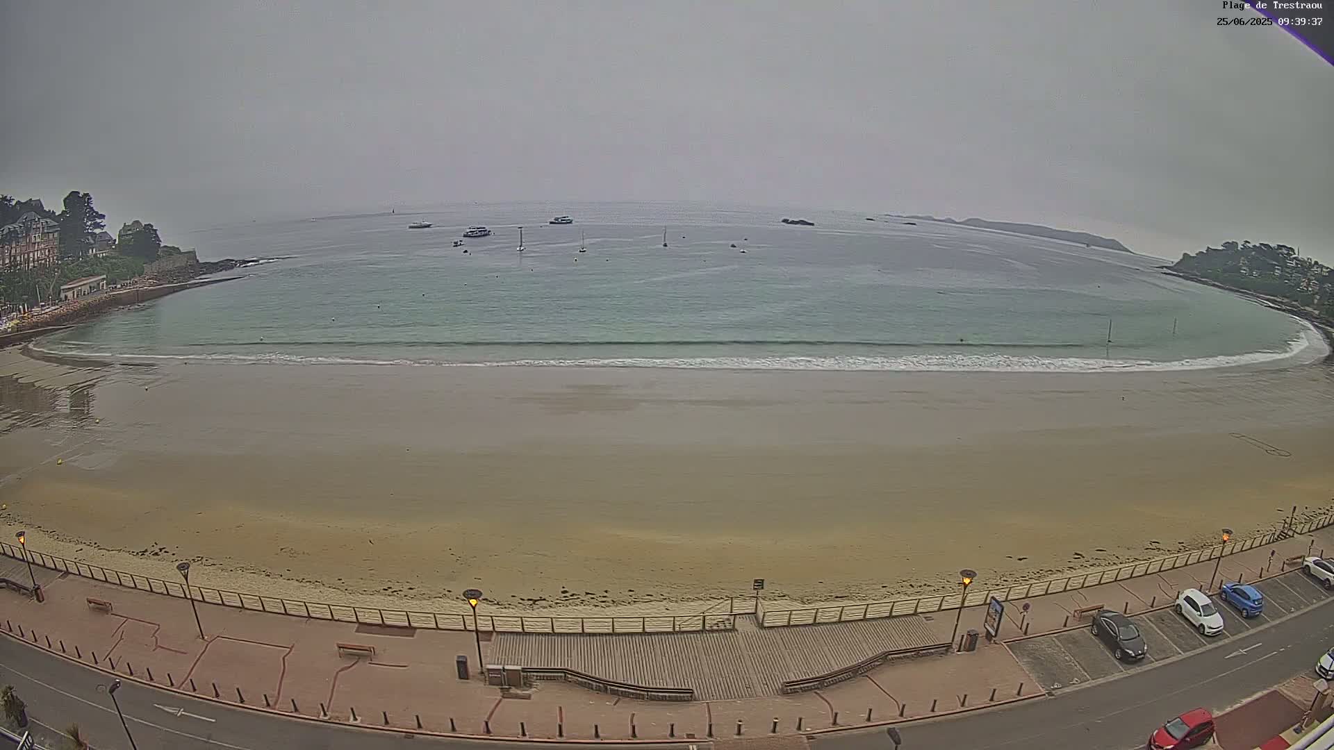 A sandy beach with small waves laps the shore under an overcast sky, with several boats visible in the calm water beyond.