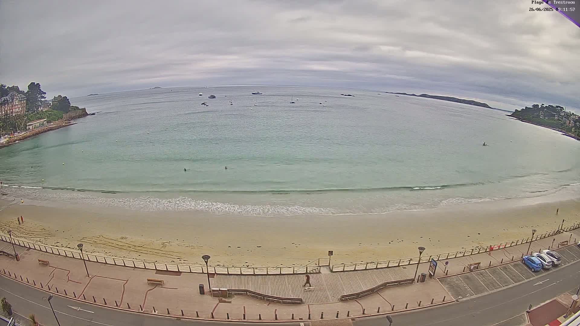A sandy beach with calm, light-green water under an overcast sky, bordered by a paved walkway, buildings, and a few parked cars.