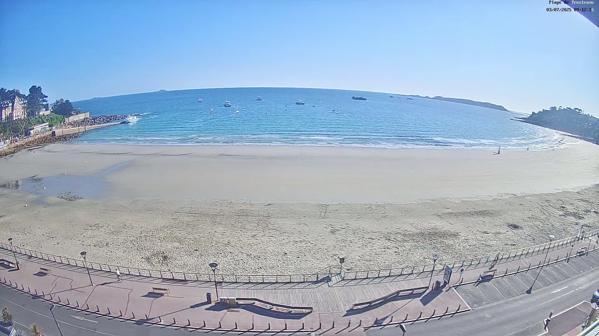 A mostly empty sandy beach stretches along a calm, light blue ocean under a clear, sunny sky.