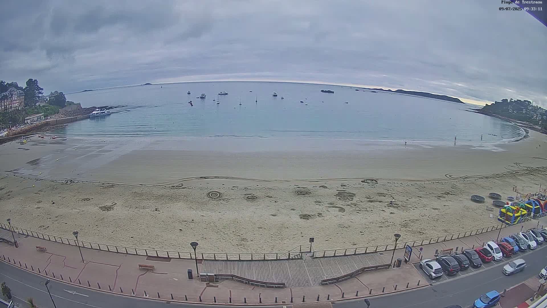 A sandy beach with several boats in the calm water beyond, under an overcast sky.