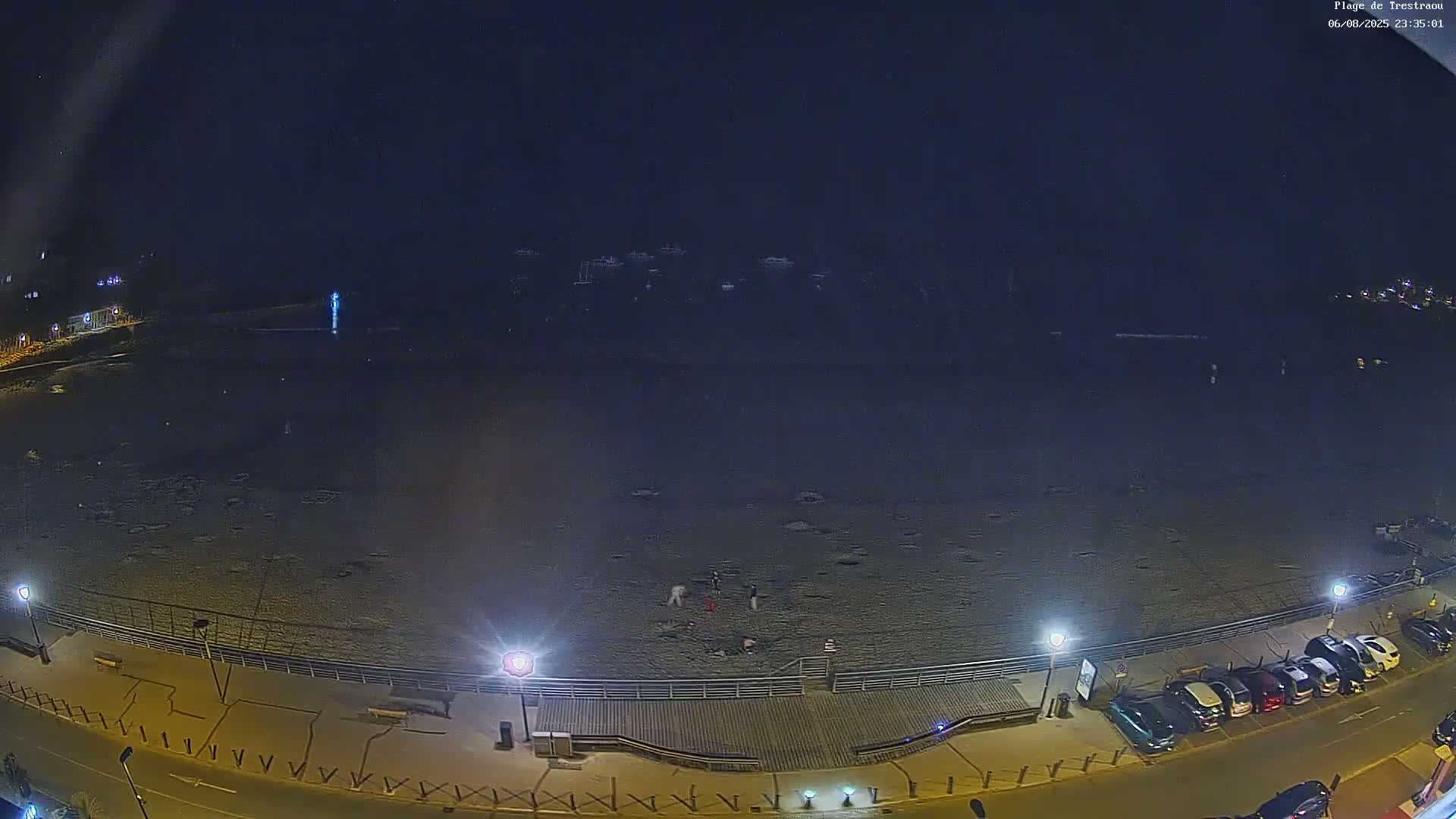 A nighttime, snow-covered beach is shown with several people gathered near a boardwalk and a row of parked cars along a street.