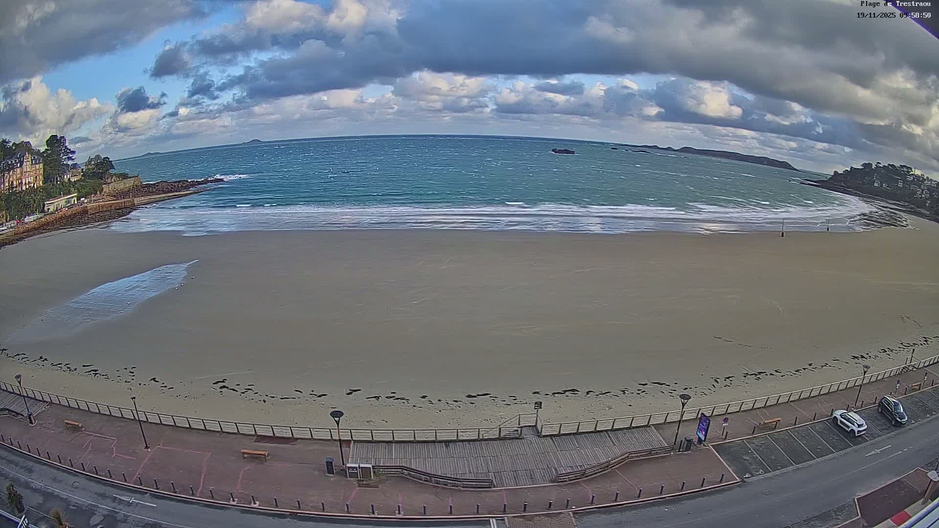 A wide sandy beach and choppy blue-green ocean with breaking waves stretch under a partly cloudy sky, bordered by a promenade with benches and parked cars, and coastal buildings nestled into a rocky shoreline.