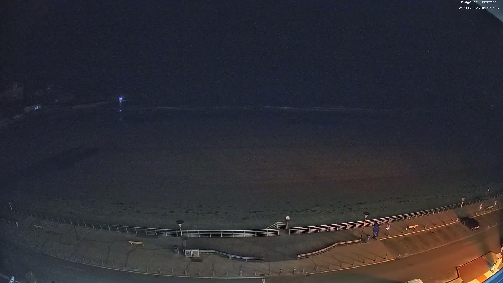 A wide sandy beach and choppy blue-green ocean with breaking waves stretch under a partly cloudy sky, bordered by a promenade with benches and parked cars, and coastal buildings nestled into a rocky shoreline.