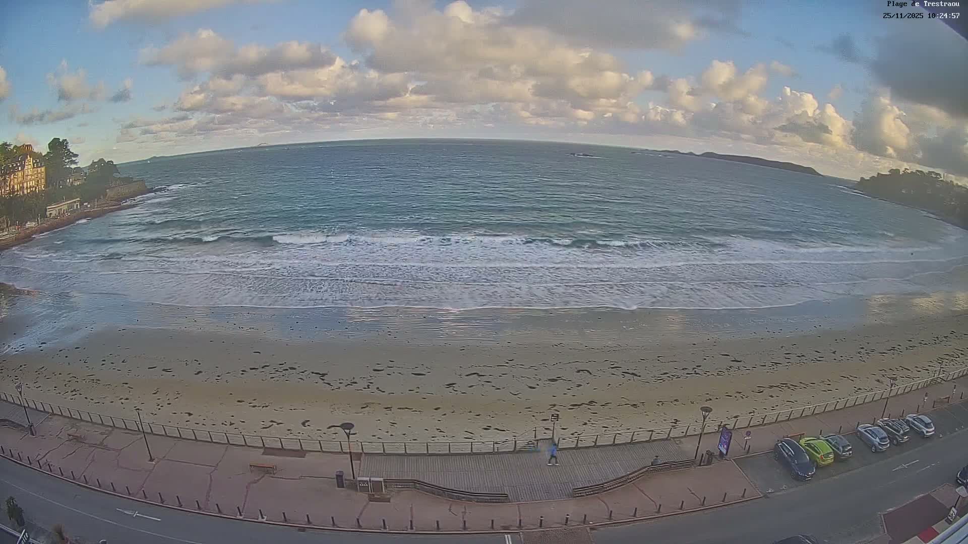 Under a partly cloudy sky with visible sunshine, a wide sandy beach curves into an ocean with gentle waves, backed by a promenade, buildings, and a street with parked cars.