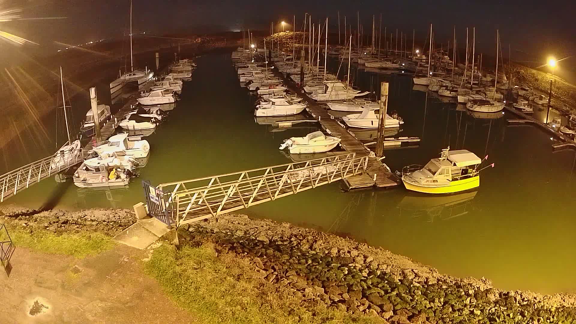 A marina at night, filled with numerous sailboats and motorboats docked along wooden piers, under dark skies.