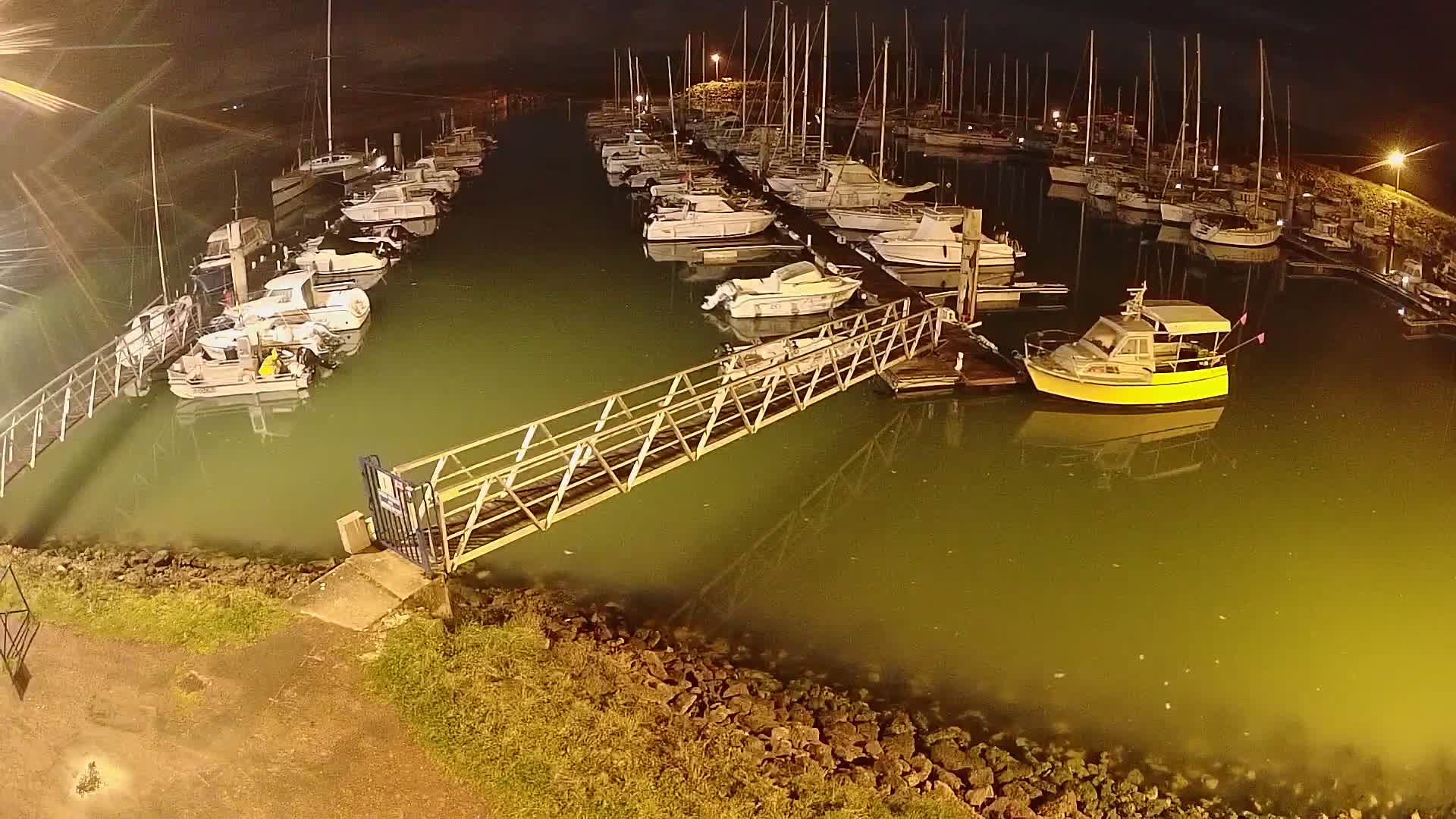 At night, a marina filled with numerous boats and yachts is illuminated, with a metal walkway extending from the shore.