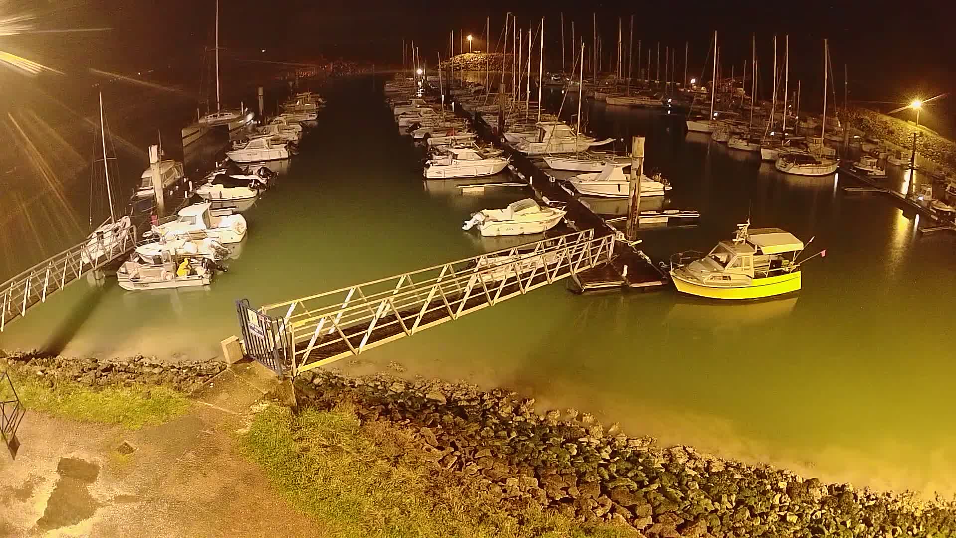 At night, a marina filled with boats is illuminated, a metal walkway connects to a dock with a yellow boat in the foreground.