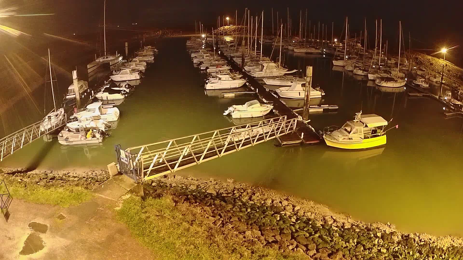 At night, a marina filled with numerous boats of varying sizes is illuminated, with a small walkway extending from the shore to the docked vessels.
