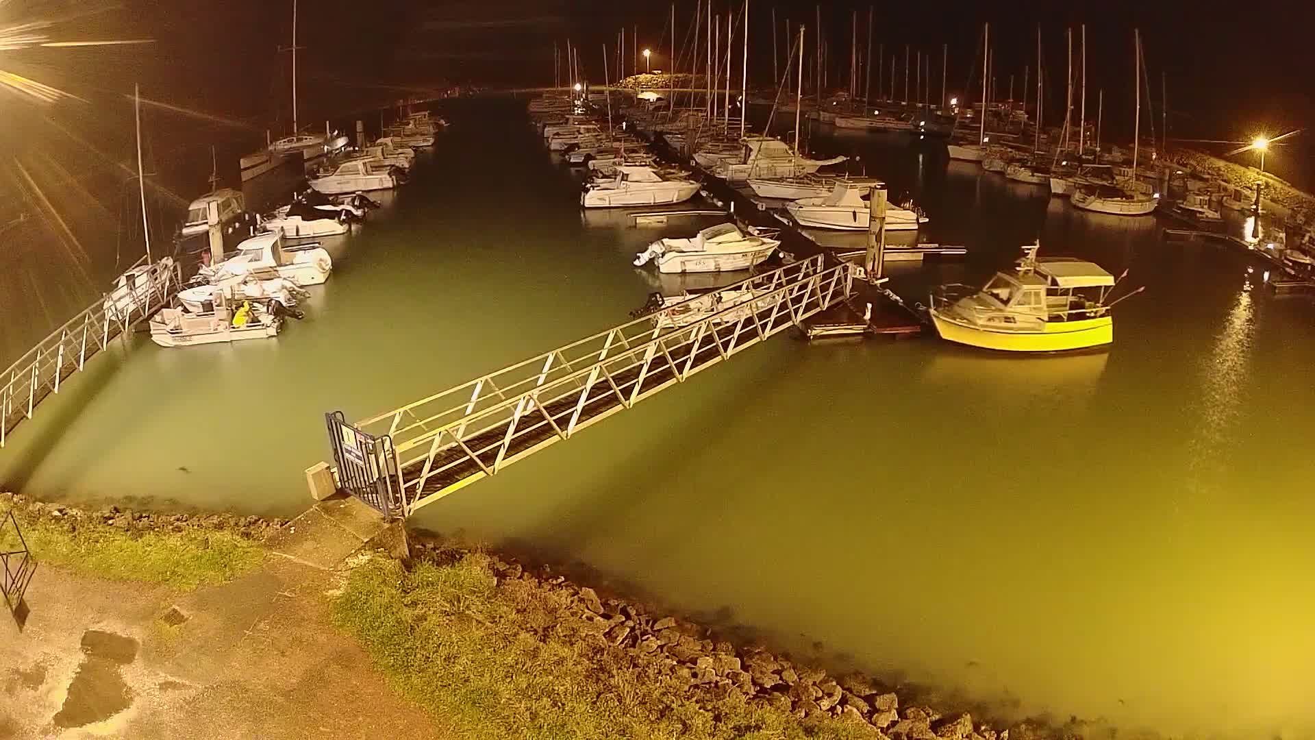 A marina at night, filled with numerous boats and yachts, is illuminated under artificial light, with a metal walkway extending from the shore.