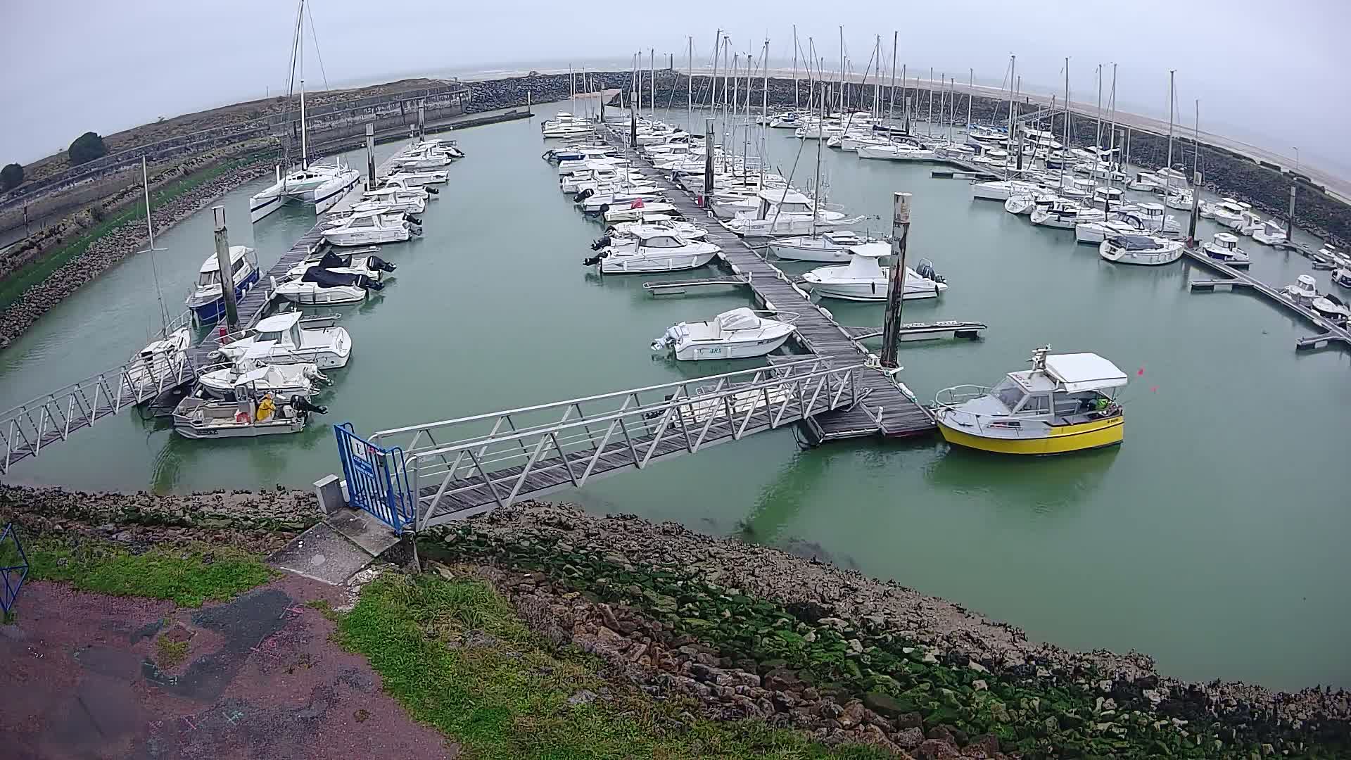 A marina filled with numerous sailboats and motorboats on a cloudy day, with a small metal walkway leading to a dock.