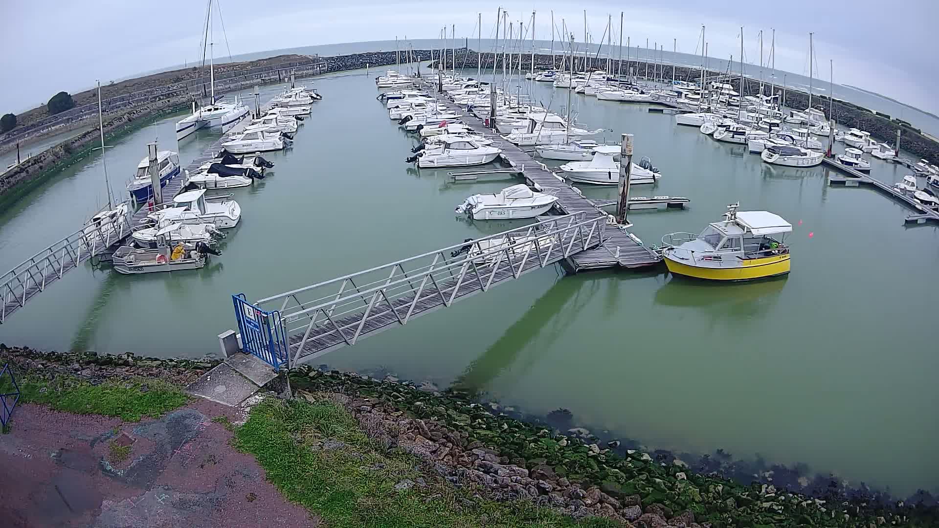 A marina filled with numerous sailboats and motorboats on a cloudy day, connected by a metal walkway to the shore.