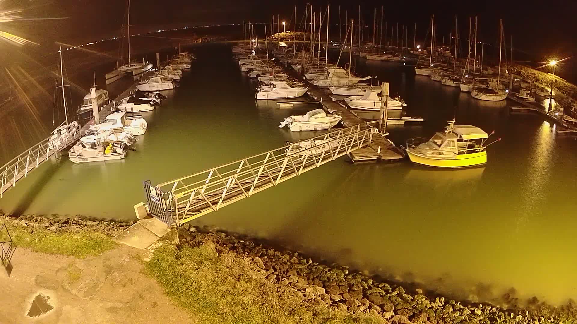 At night, a marina filled with numerous boats and yachts is seen from above, with a metal gangway leading to a dock.