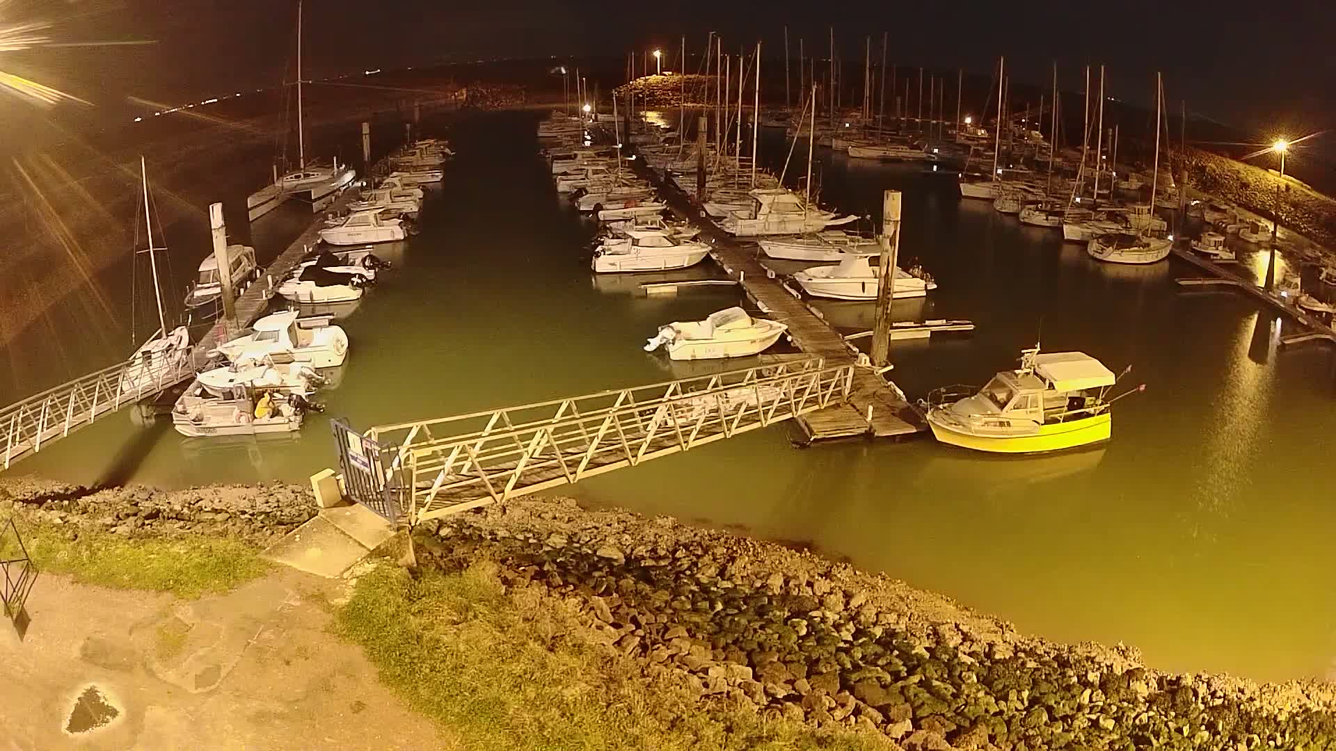 At night, numerous sailboats and motorboats are docked in a marina with calm, greenish water, illuminated by artificial light.