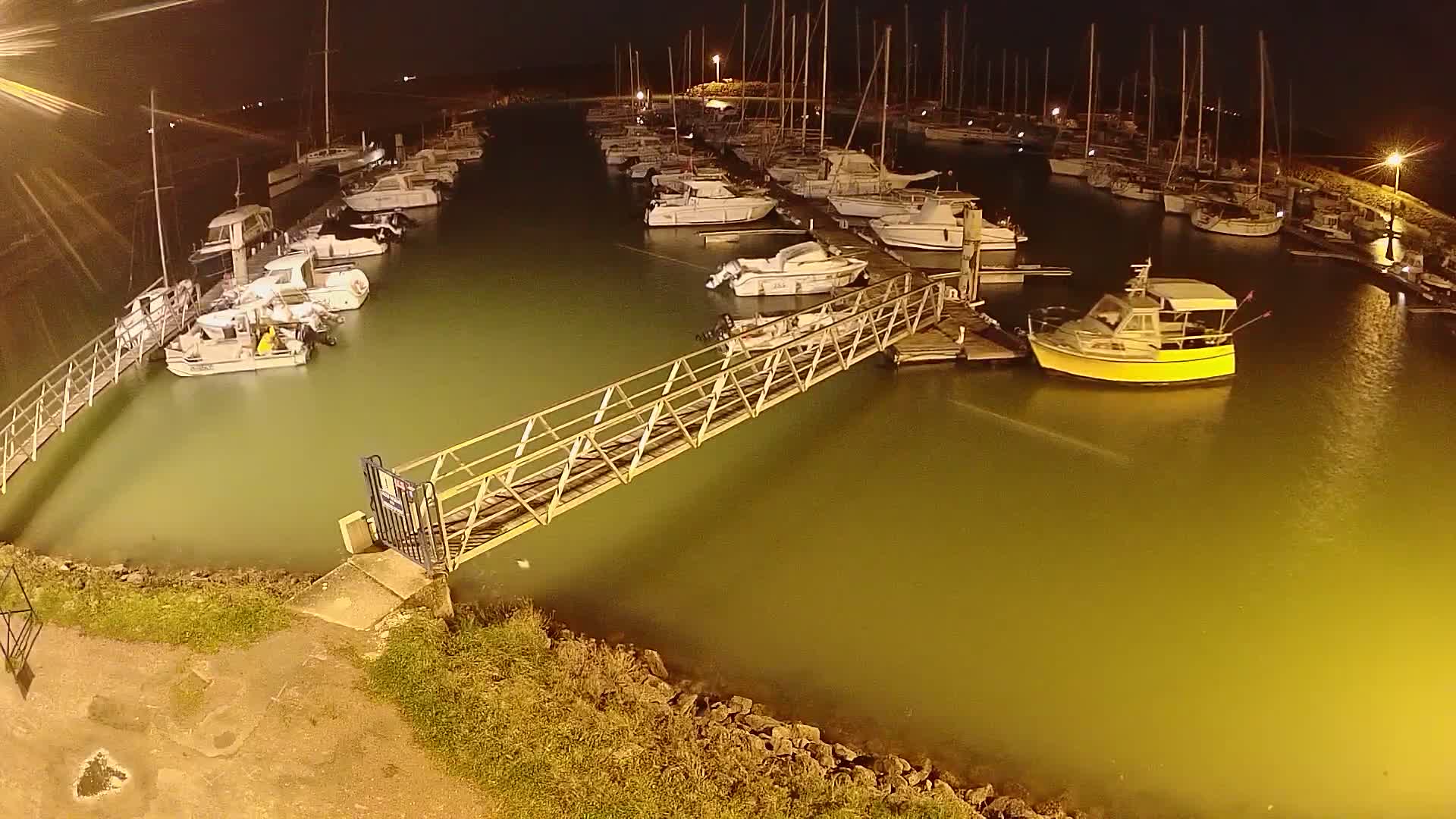 A marina at night, illuminated by artificial lights, shows numerous sailboats and motorboats moored along docks connected by a metal walkway, with the water appearing a murky greenish-yellow.